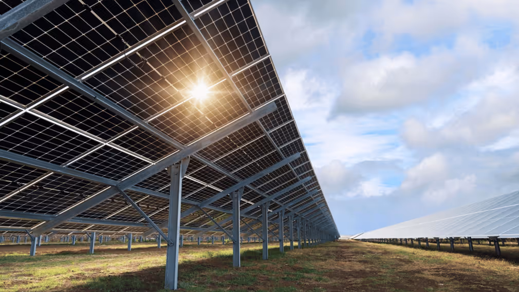 Rows of solar panels installed in a field under a partly cloudy sky with sunlight shining through one panel.