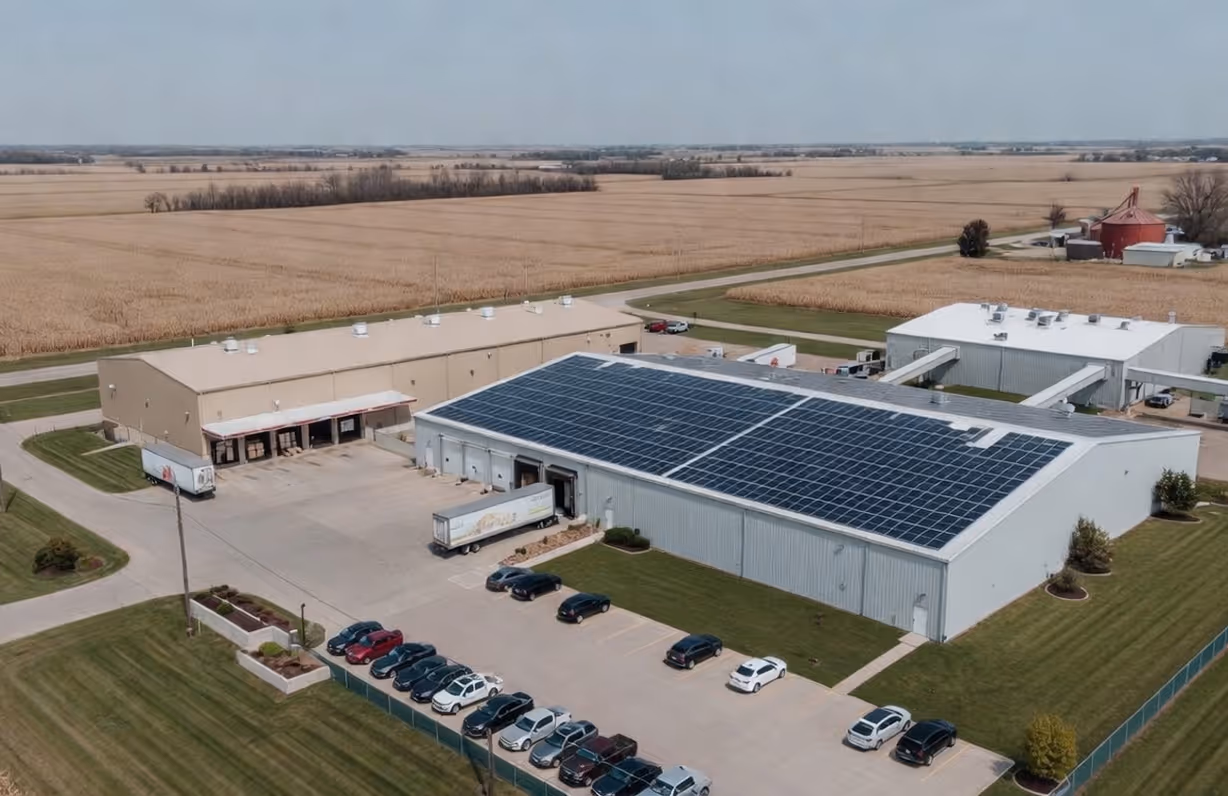 Aerial view of an industrial complex with multiple buildings, one with solar panels on the roof, surrounded by farmland and parked cars.