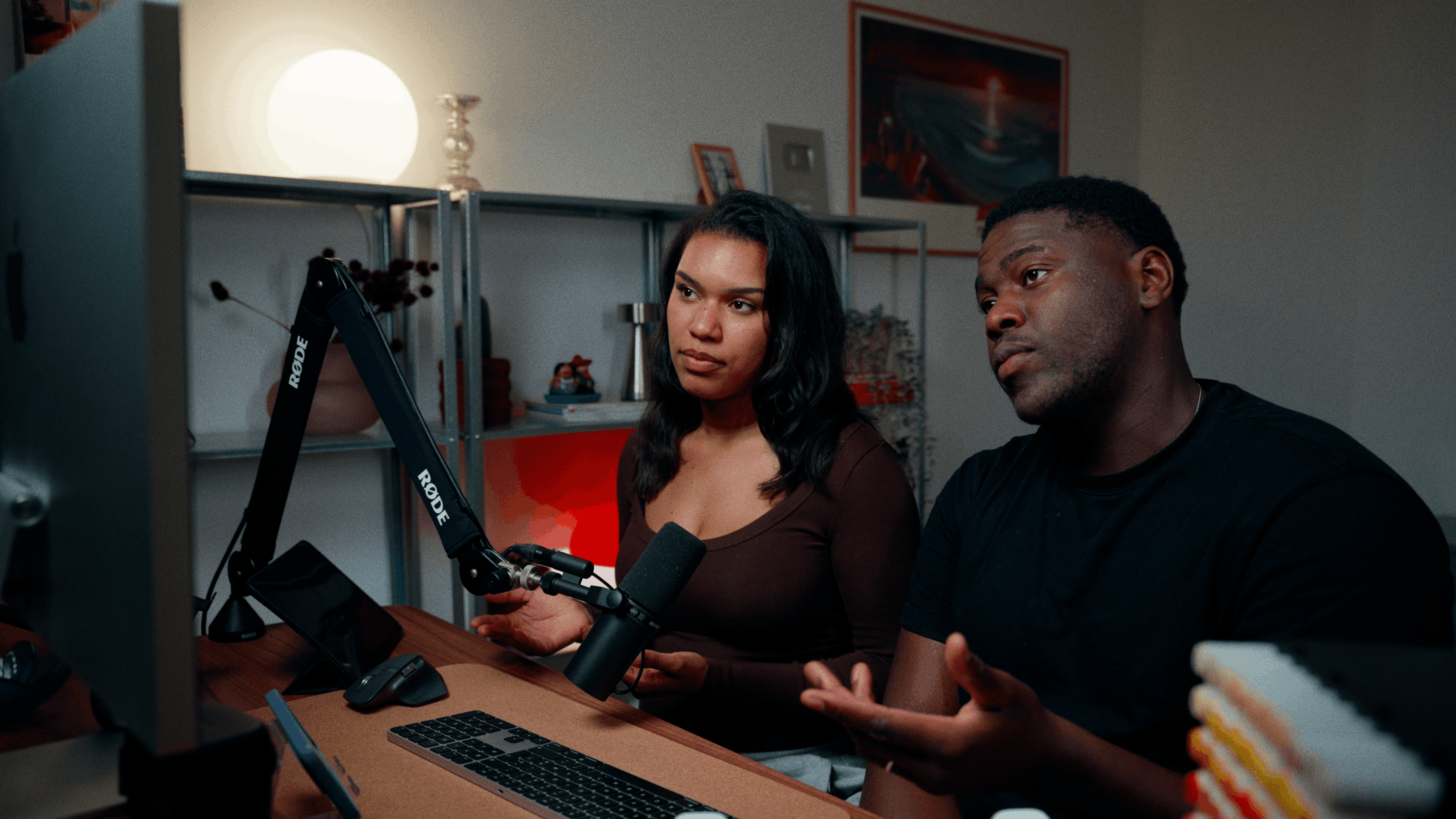Man wearing a baseball cap working at a desk with an Apple computer and using a microphone in a dimly lit room.