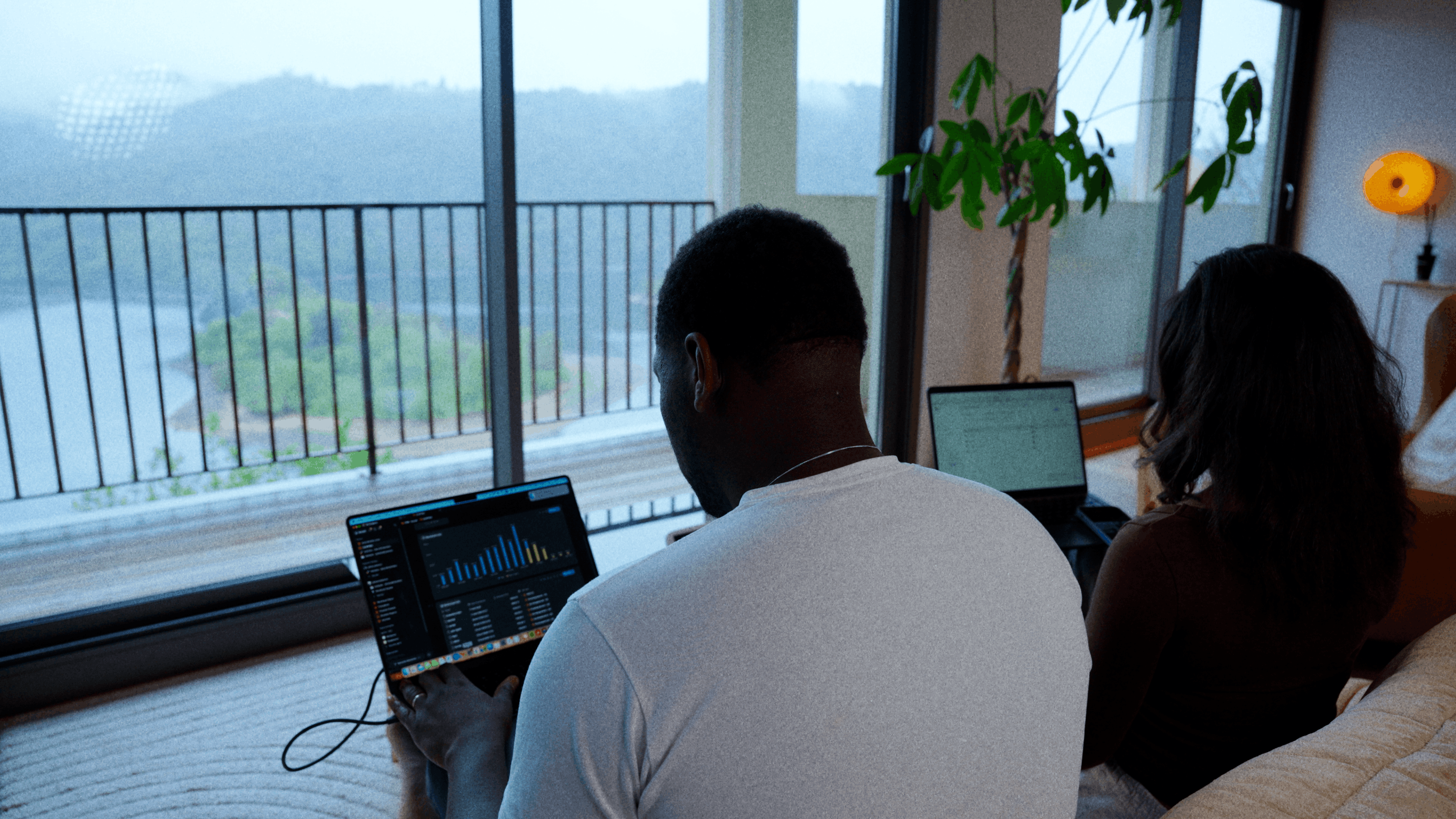 Man wearing a gray beanie and jacket standing by a calm lake with mountains and trees in the background.