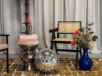 Moderne zithoek met zwarte stoelen met gevlochten rugleuningen, een metallic bijzettafel met boeken en een rode kaars, een discobal op een bruingeblokt tapijt en een vaas met kleurrijke bloemen.