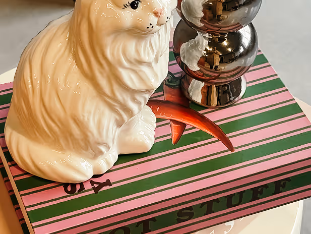 Witte keramische katbeeldje en een glanzende metalen kandelaar met drie bollen op een gestreept roze en groen boek.