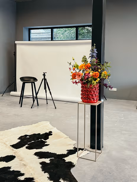Binnenruimte met een zwart-wit koeienhuid op de vloer, een hoge rode vaas met kleurrijke bloemen op een metalen standaard, een zwarte kruk, een camerastatief en een witte fotobackdrop tegen een grijze muur.
