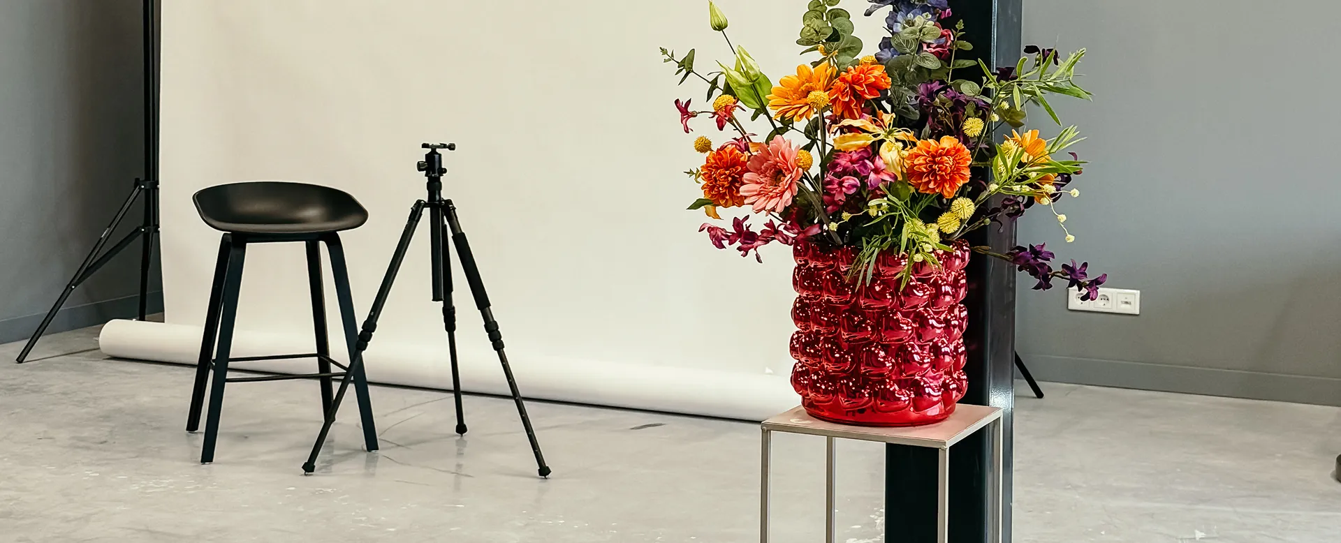 Een kleurrijk boeket bloemen in een rode vaas op een metalen tafeltje in een fotostudio met een zwarte kruk en een statief.