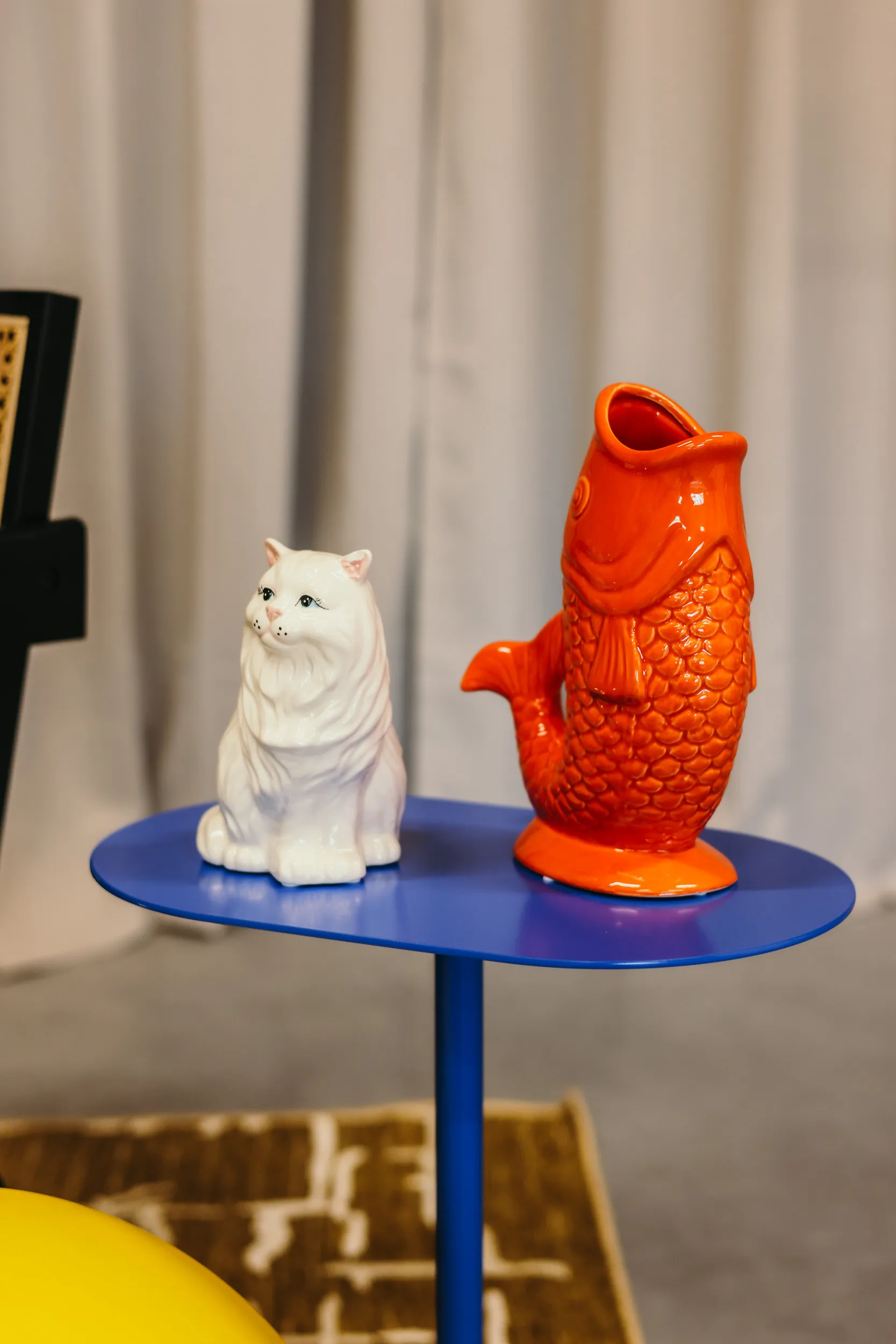 Een wit keramisch beeld van een zittende kat naast een oranje visvormige kan op een blauw tafeltje.
