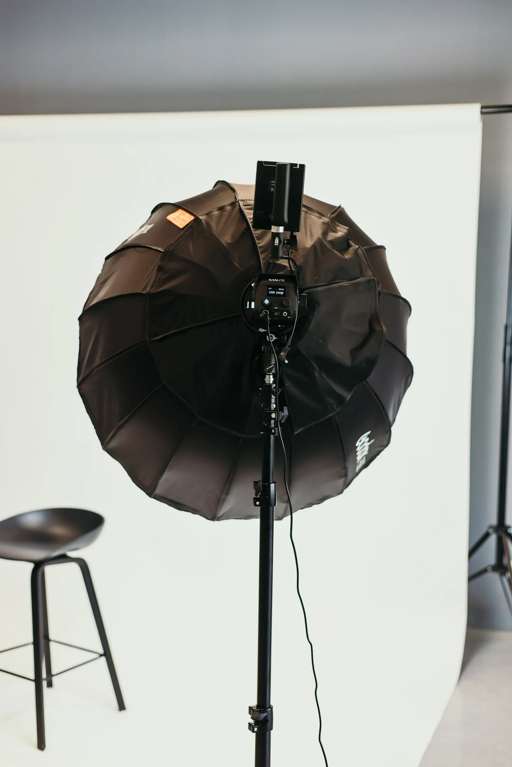 Zwarte studiofotolamp met paraplu diffuser voor een witte achtergrond, naast een zwart krukje.