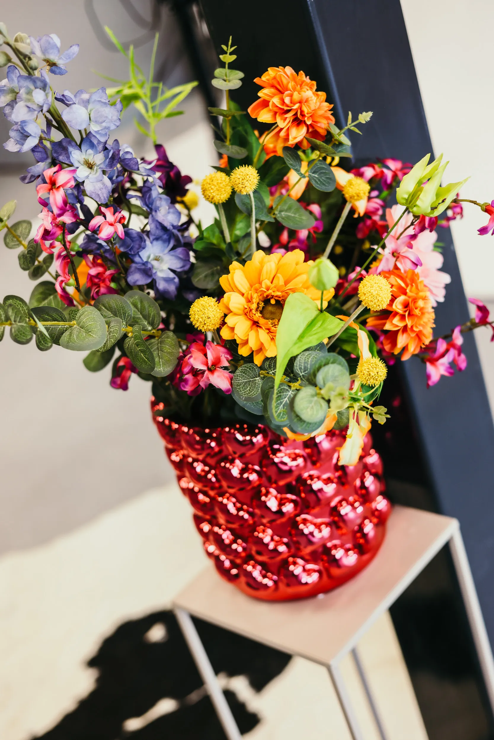 Een boeket kleurrijke bloemen in een glanzende rode vaas geplaatst op een klein tafeltje.