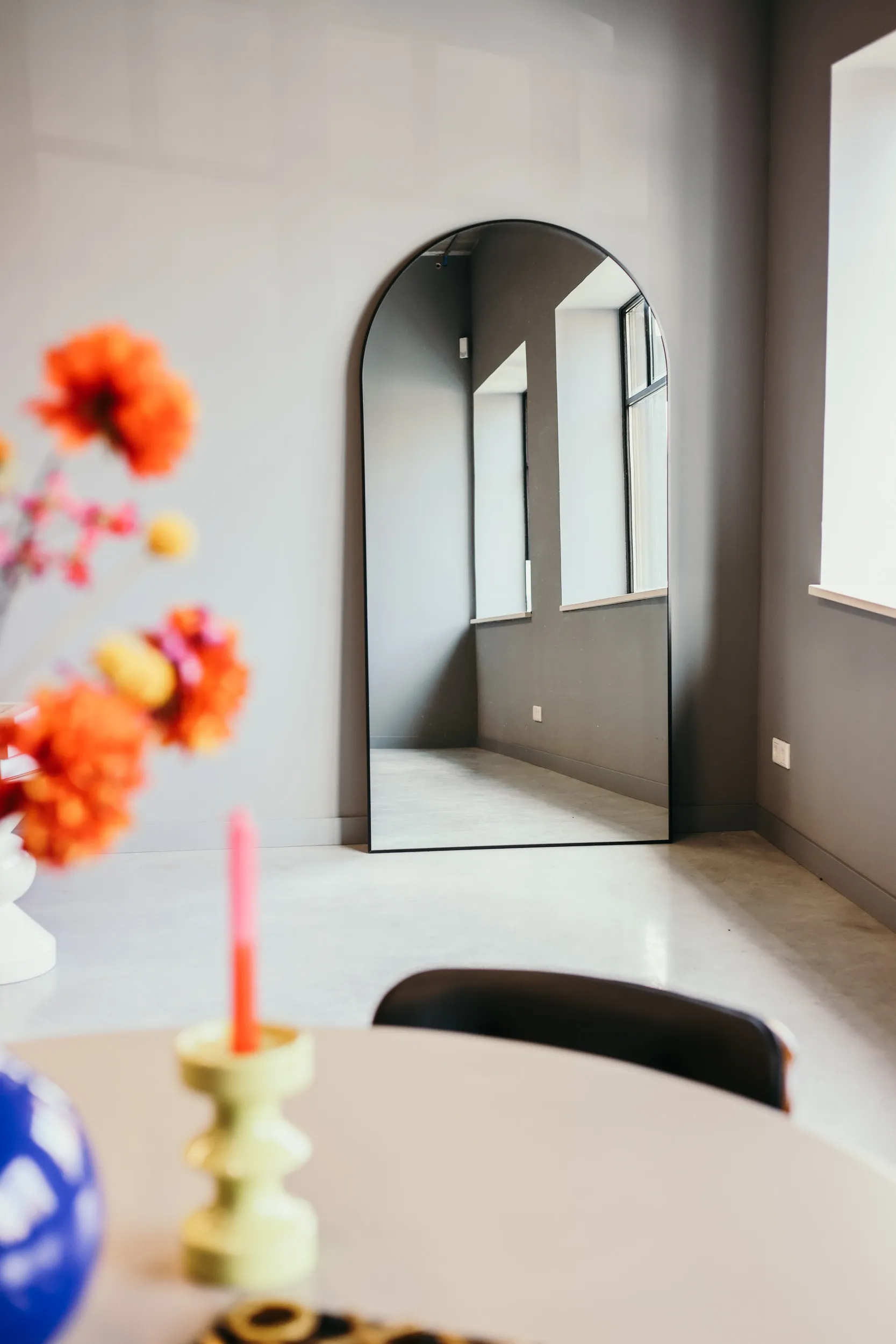 Grote, boogvormige spiegel met zwarte rand tegen een grijze muur, met vaas met oranje en gele bloemen en kaars op tafel op de voorgrond.