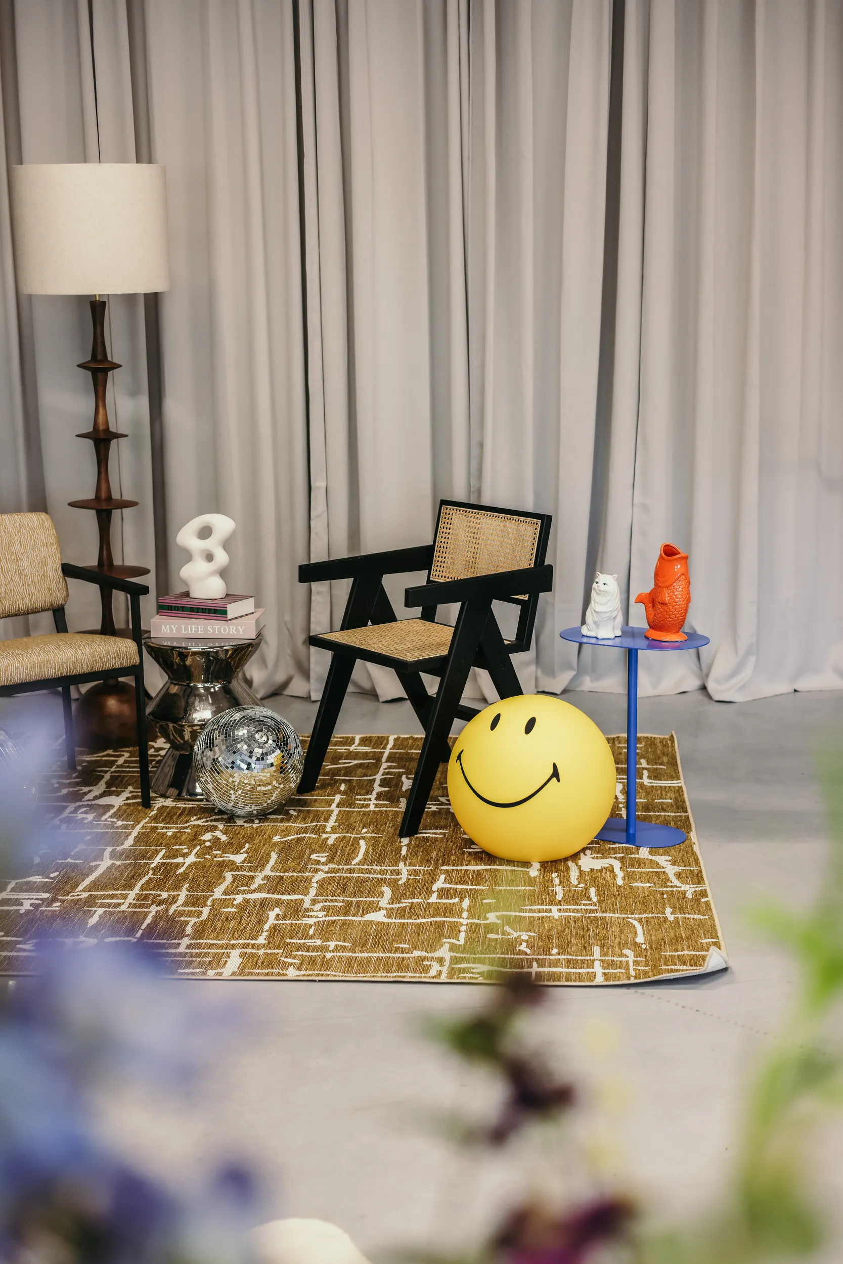 Gezellige woonkamerhoek met twee stoelen, een bruine geometrische vloerkleed, een staande lamp, een disco bal, en een gele smiley bal.