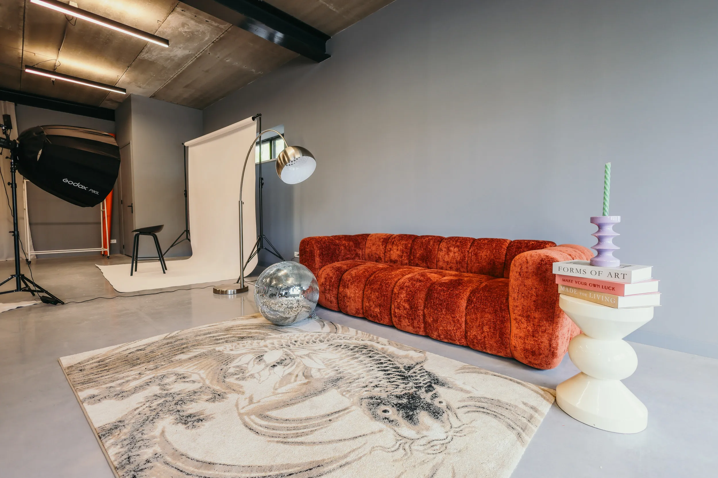 Moderne woonkamer met een oranje fluwelen bank, sierlijke karper-print vloerkleed, discobal, en minimalistisch bijzettafeltje met kaars en boeken.