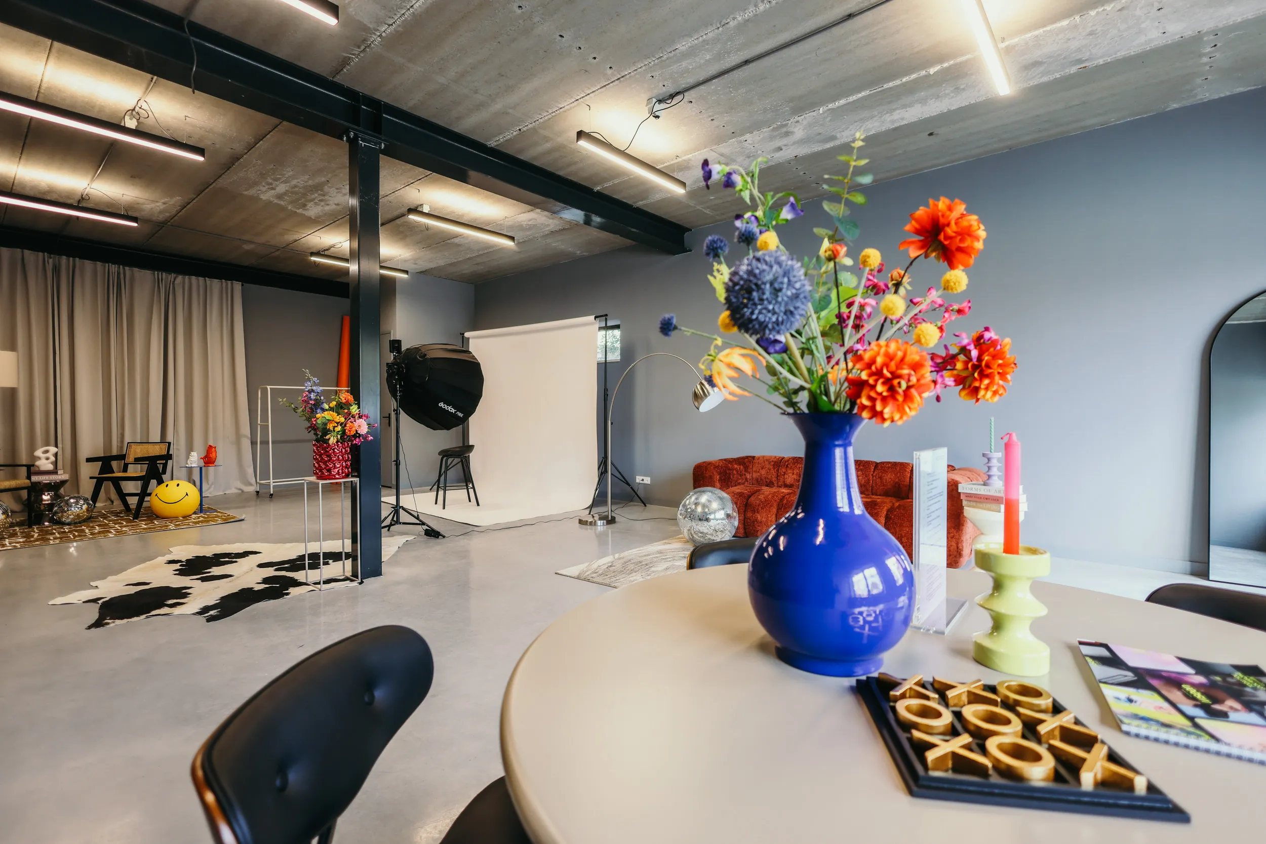 Interieur van een moderne studio met een ronde tafel met een blauwe vaas met kleurrijke bloemen, een groene kaarshouder, een tic-tac-toe spel en stoelen.