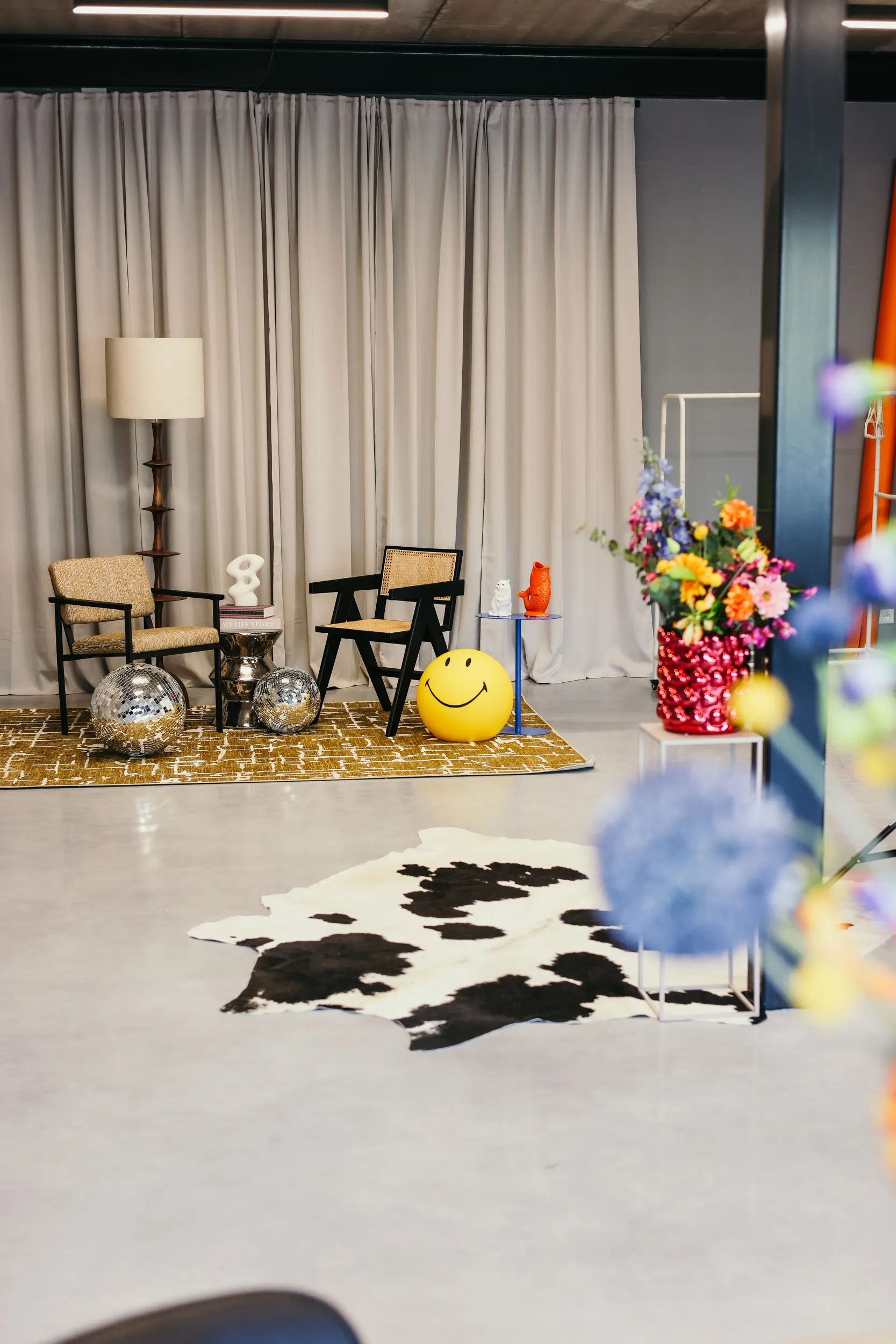 Moderne zithoek met twee stoelen, een okergeel vloerkleed, een staande lamp, discoballen, een gele smileybal, een blauwe bijzettafel en een vaas met kleurrijke bloemen.