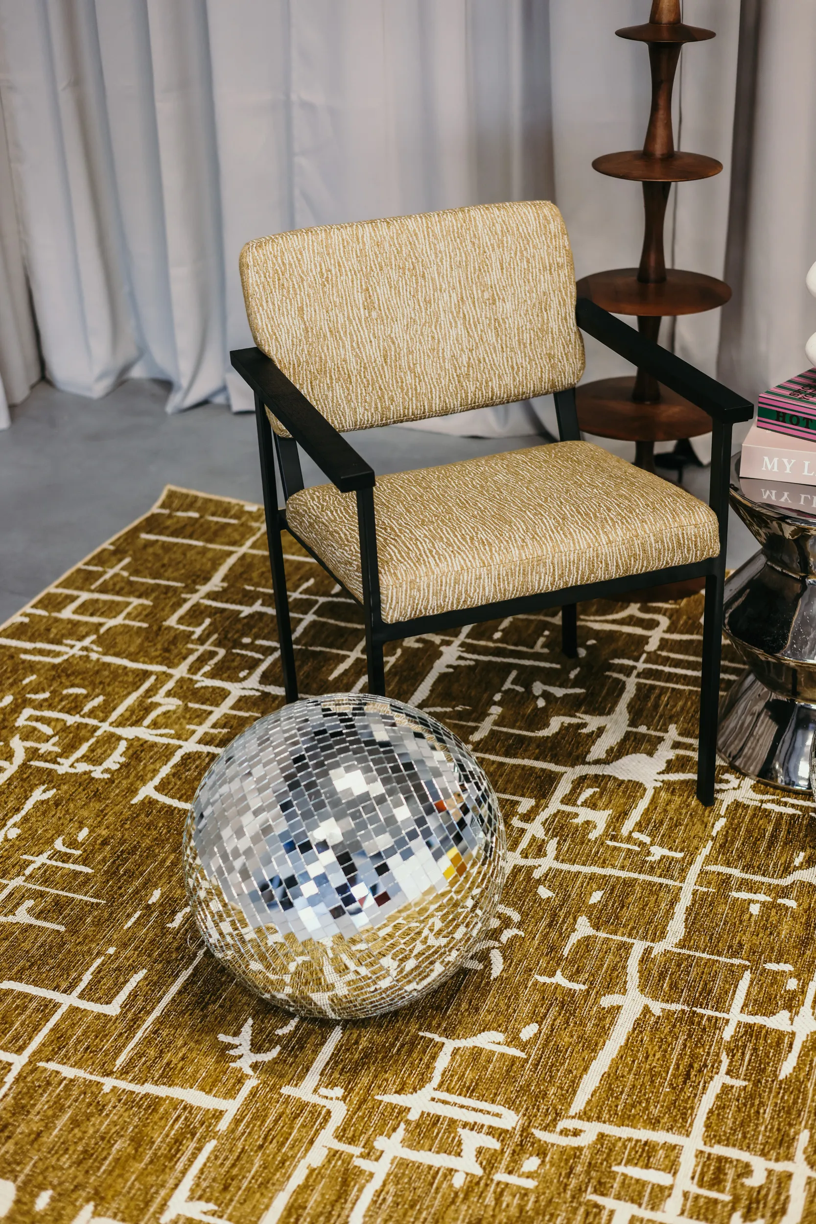 Modern beige armchair met zwarte armleuningen op een okergeel tapijt met wit grafisch patroon, naast een glimmende discobal.