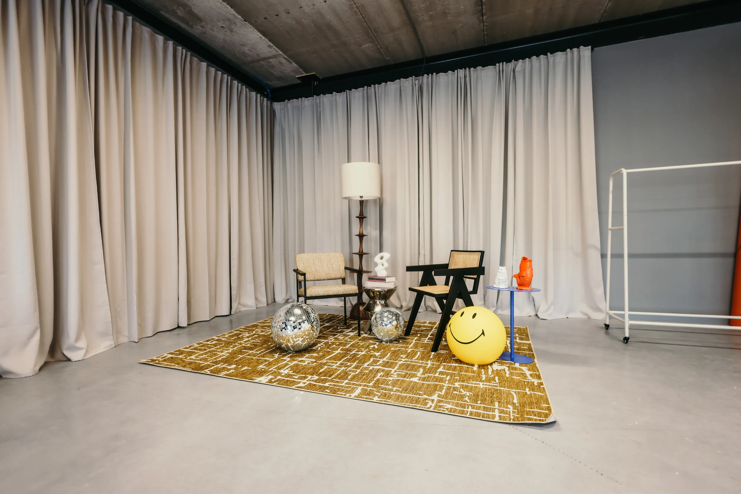 Moderne kamer met twee stoelen, een staande lamp, disco ballen, een geel gelukkig gezicht bal, blauwe bijzettafel met decoraties, en een geel patroon tapijt, omringd door grijze gordijnen.