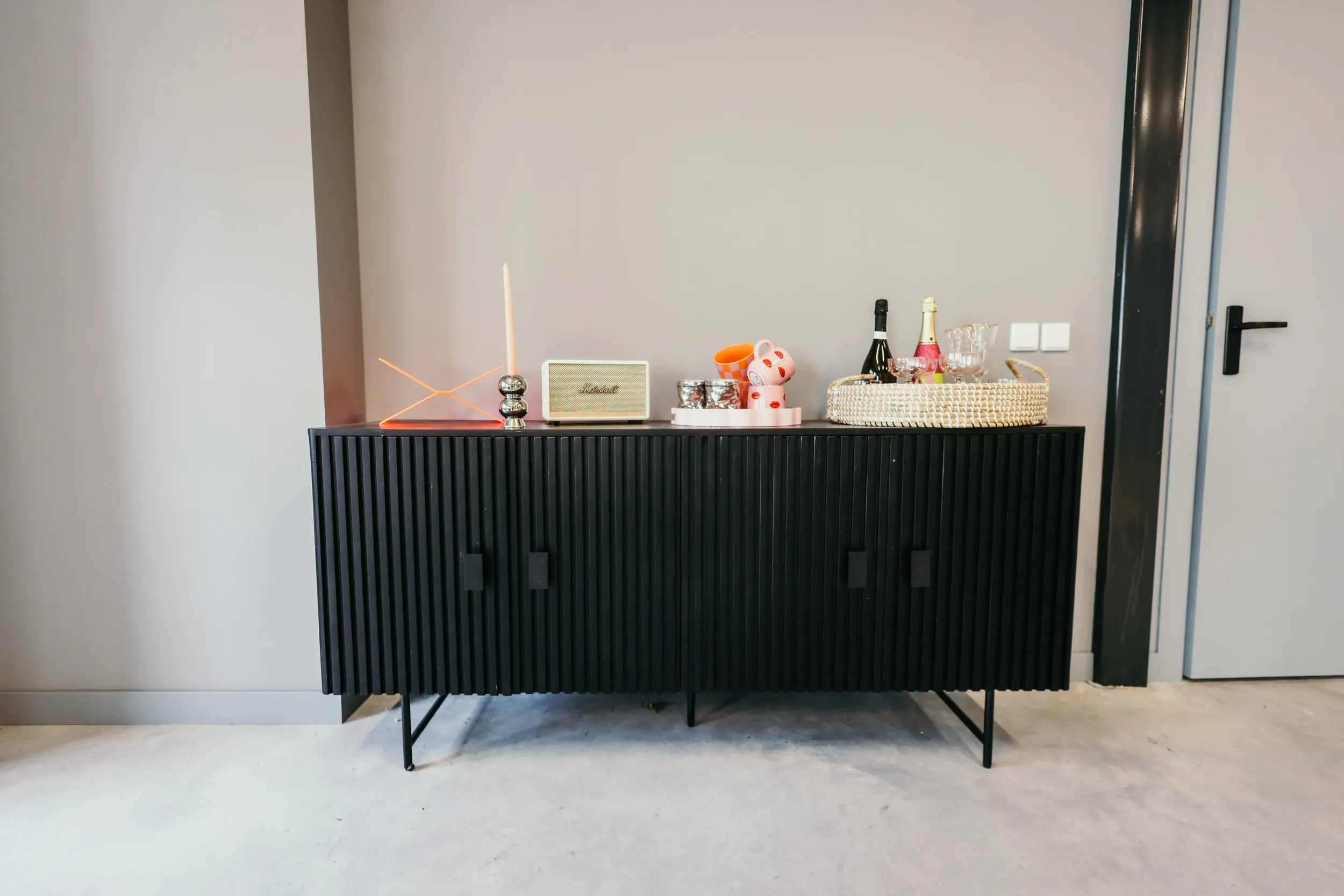 Zwarte kast met decoratieve voorwerpen waaronder een kaars, luidspreker, decoratie en een mand met flessen en glazen.