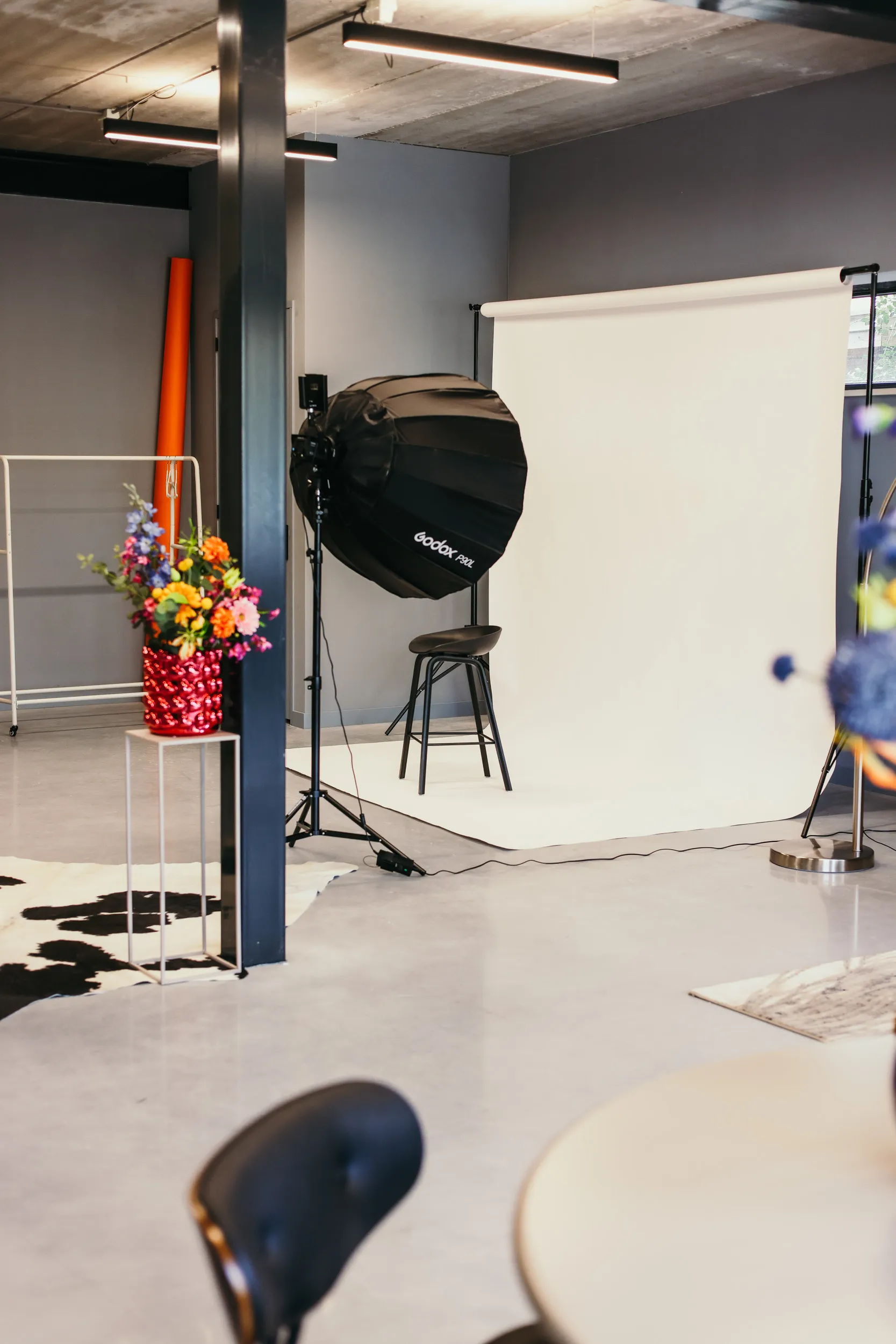 Fotostudio met een zwarte stoel, witte achtergrond, Godox softboxlamp en een vaas met kleurrijke bloemen.