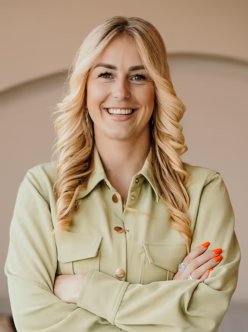 Vrouw met lang, krullend blond haar en oranje nagellak, glimlachend met armen over elkaar in een groene blouse.