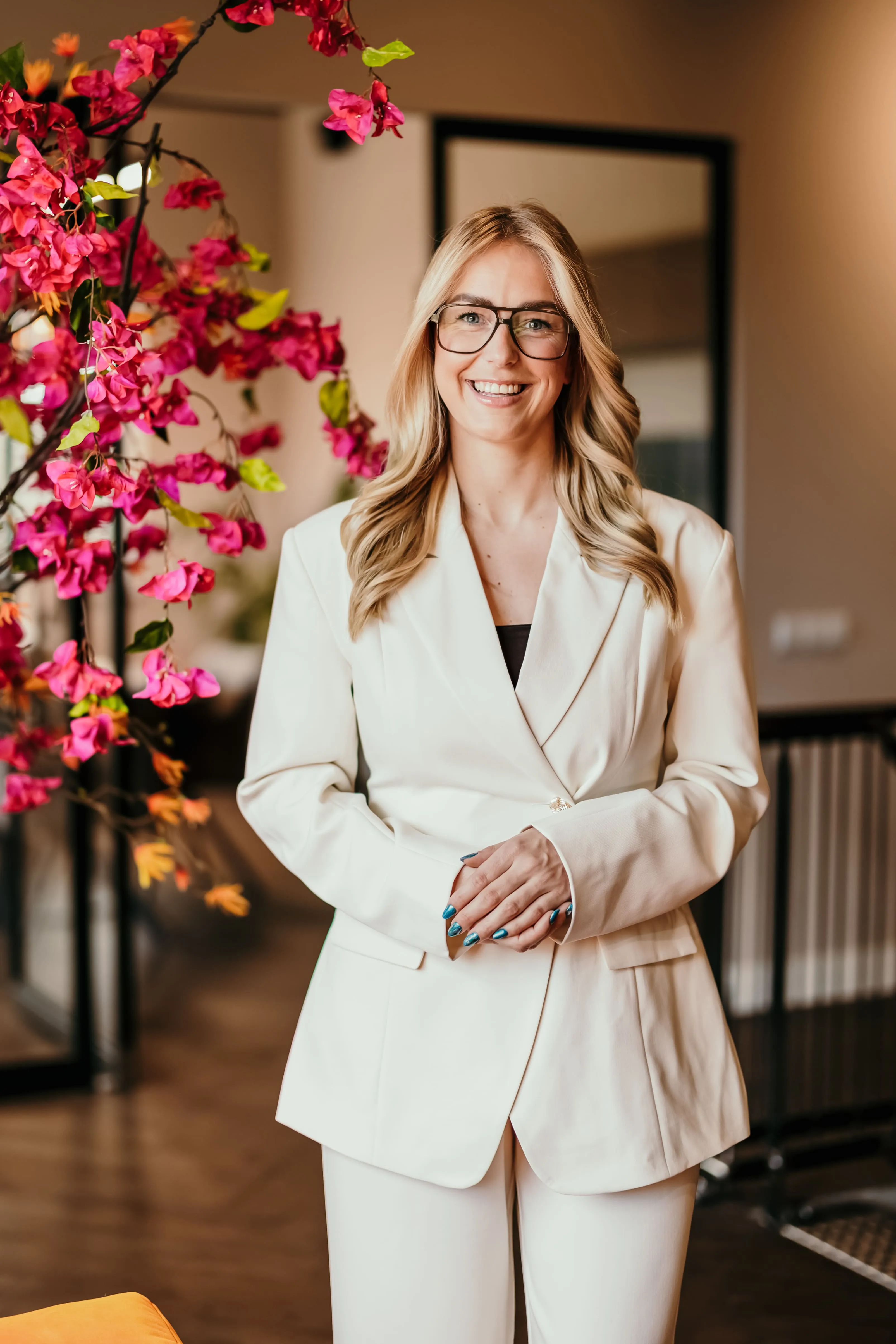 Vrouw met blonde golvende haren en bril, gekleed in een witte blazer en broek, glimlachend in een binnenruimte met roze bloemen op de achtergrond.