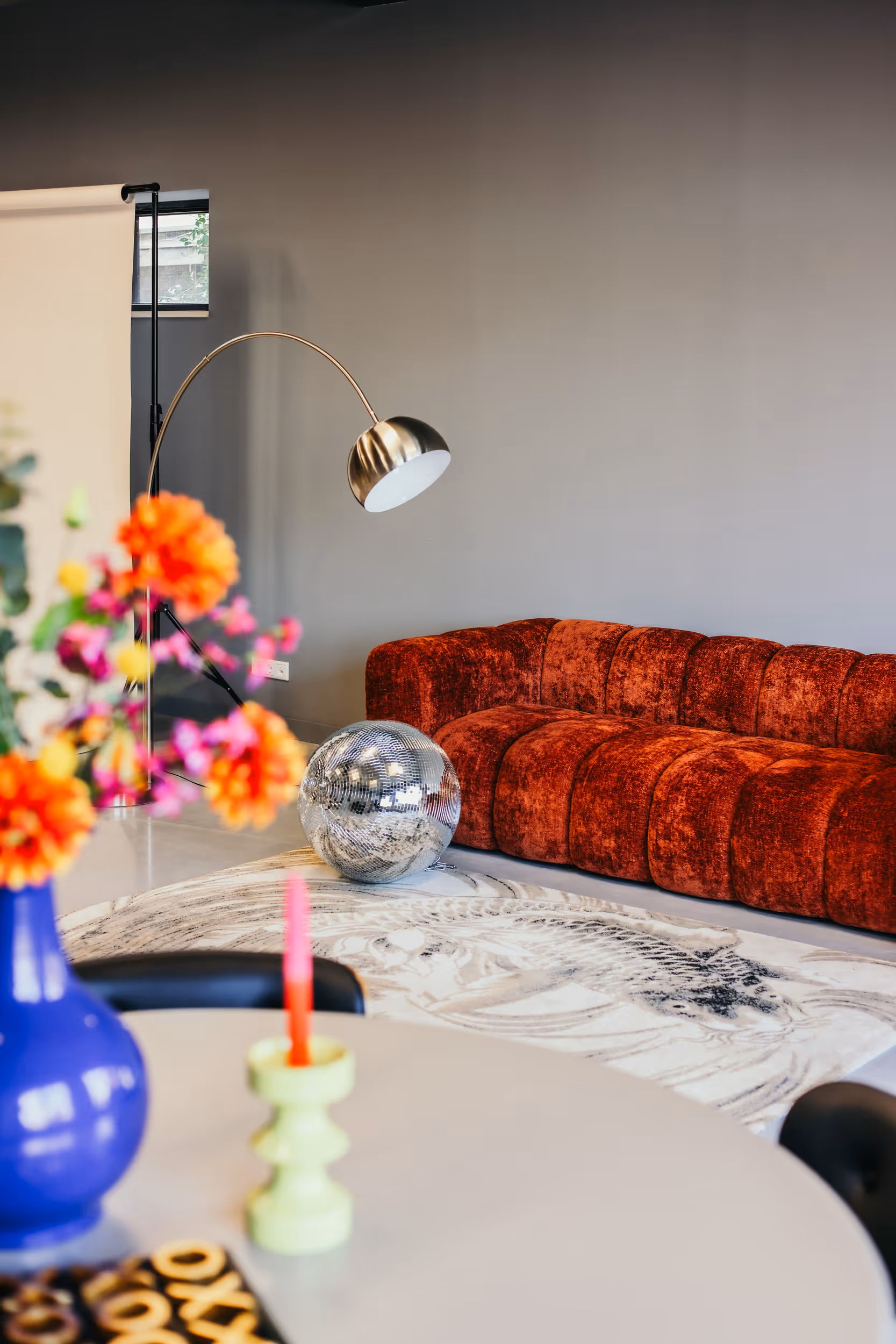 Moderne woonkamer met een pluche, roodbruine bank, een zilveren discobal op een gedetailleerd tapijt en een booglamp tegen een grijze muur.