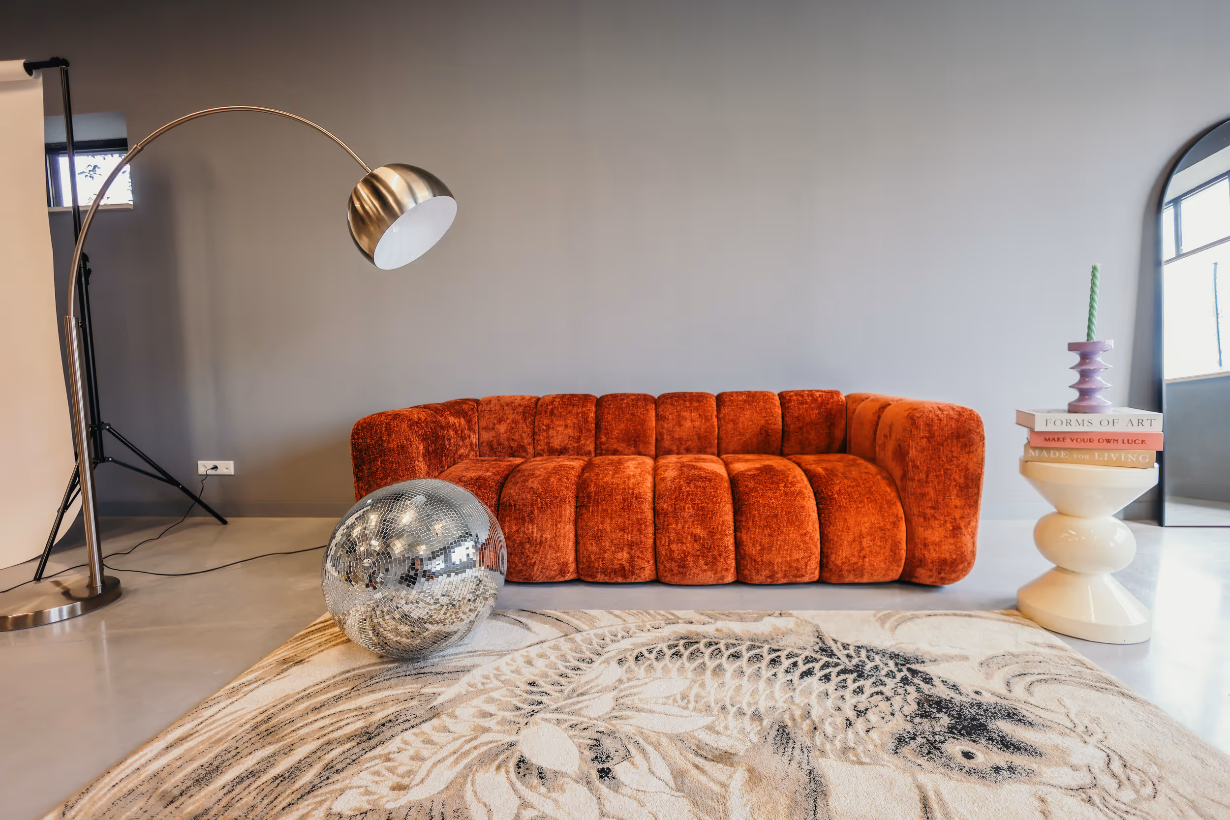Moderne woonkamer met een oranje fluwelen bank, een grote zilveren bol, een staande lamp en een wit bijzettafeltje met boeken en kaars.