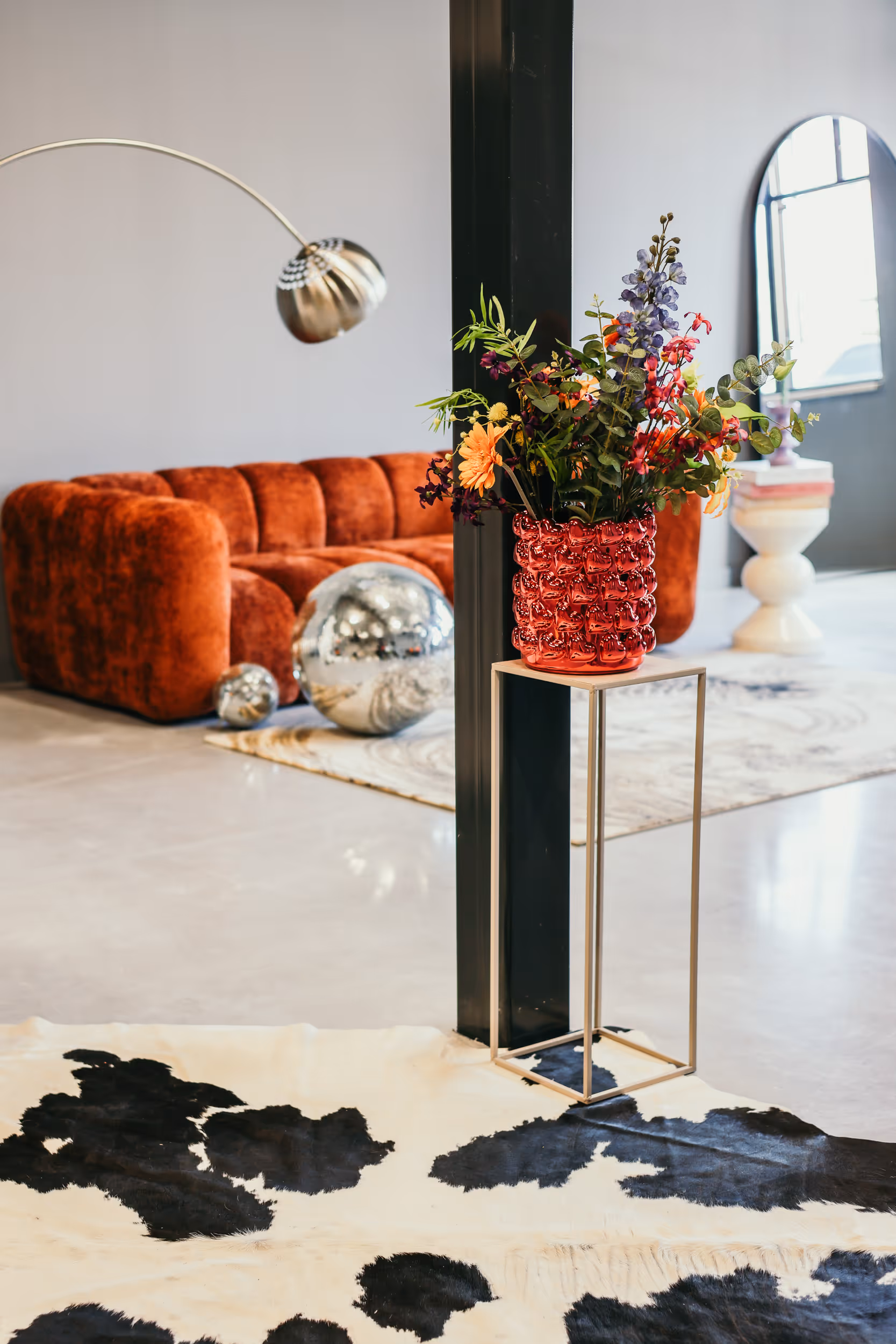 Moderne woonkamer met een oranje fluwelen bank, zilveren decoratieve bollen, een bloemstuk in een rode vaas op een metalen standaard en een zwart-witte koeienhuid tapijt.
