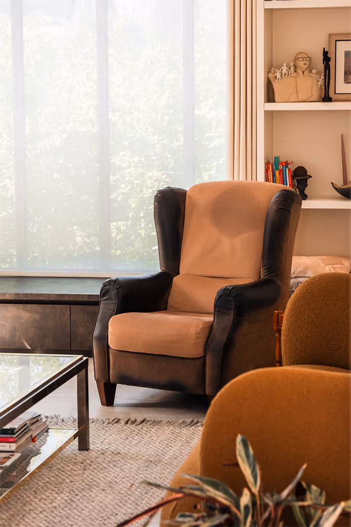 Gezellige woonkamer met een bruine fauteuil, een glazen salontafel, een boekenkast en een groot raam met vitrages.