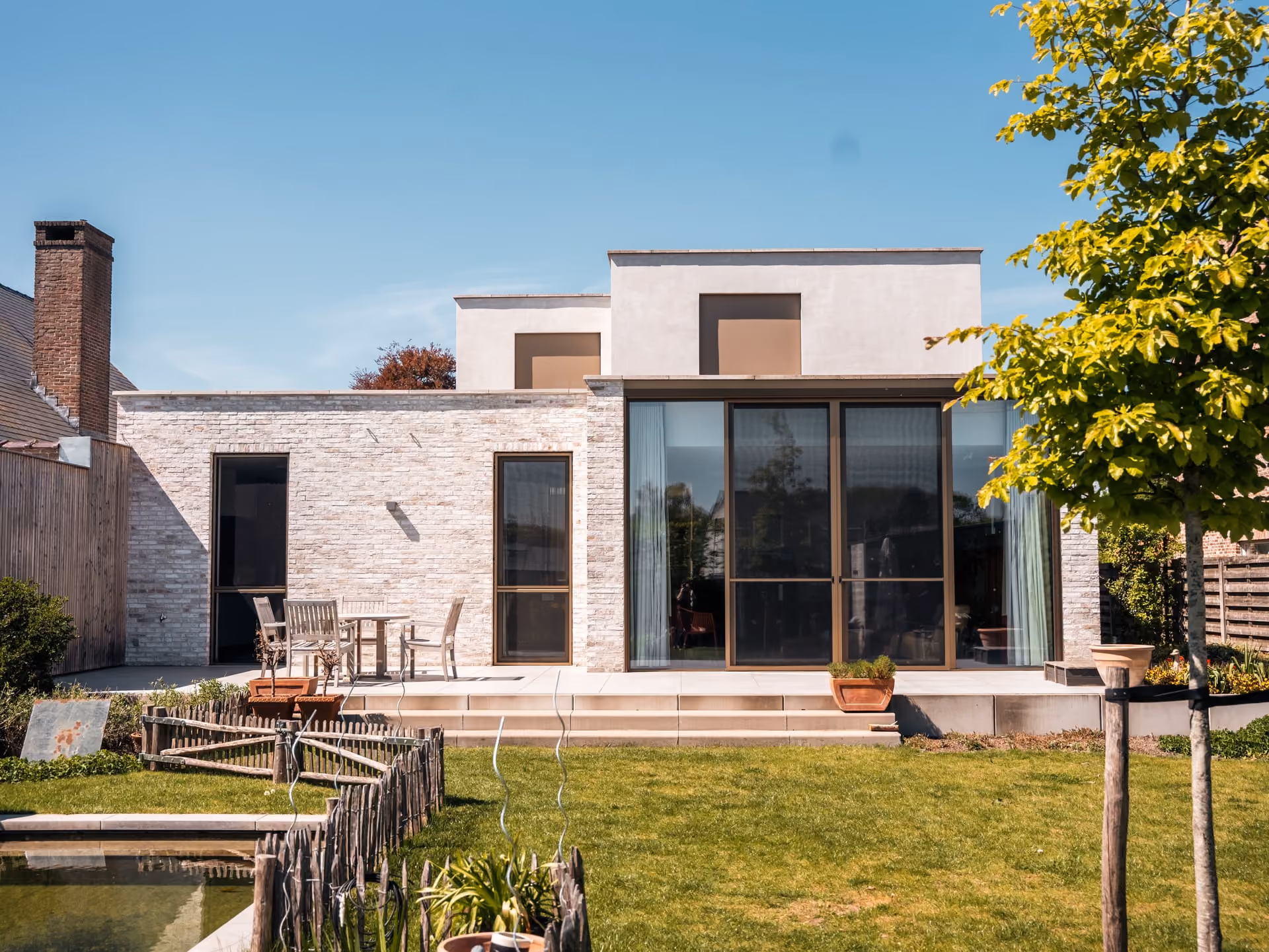 Modern huis met grote glazen deuren, terrasmeubilair, een tuin en een kleine, omheinde vijver.