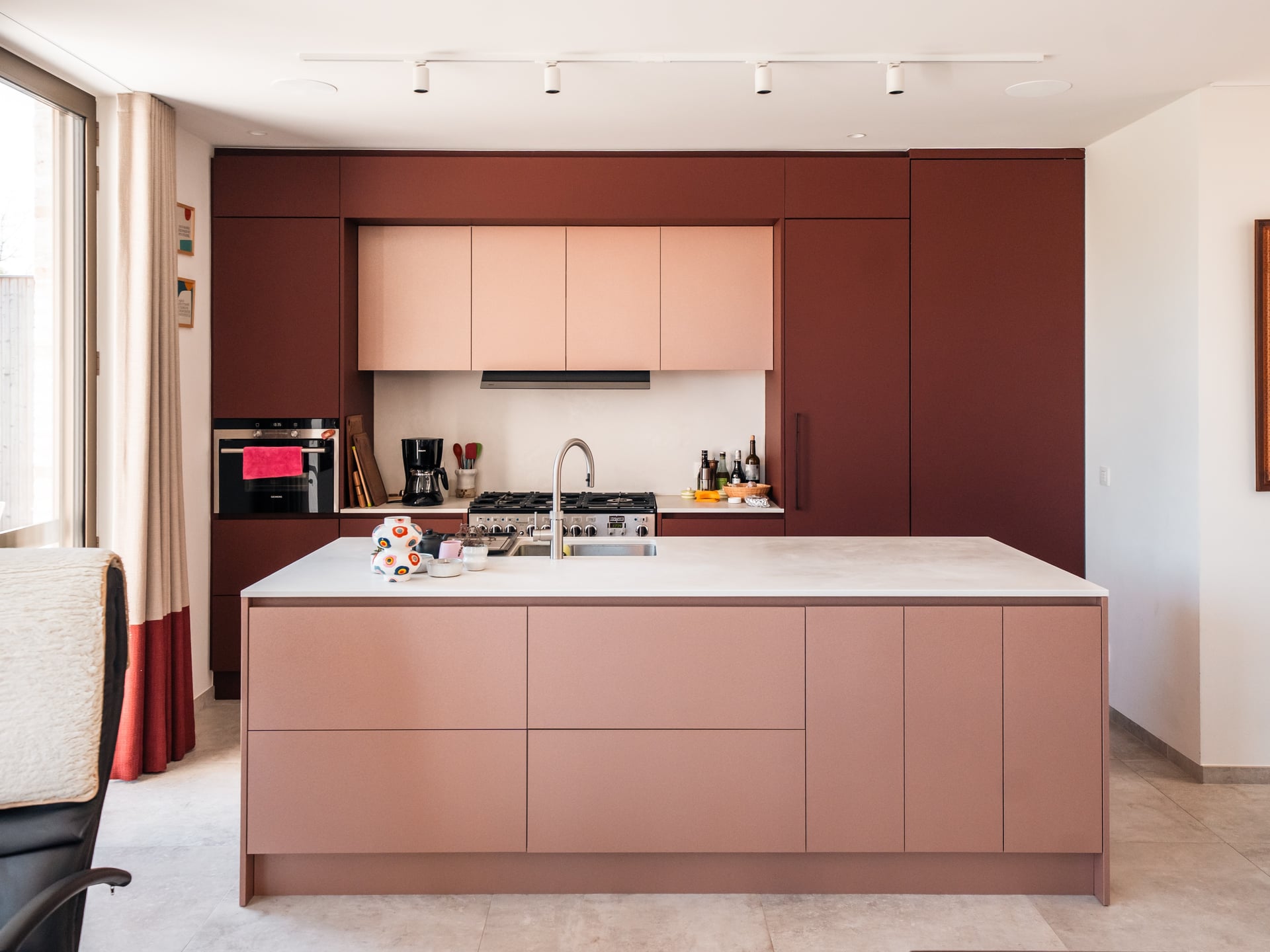Moderne keuken met roodbruine kastwanden, roze eiland en gasfornuis in een lichte open leefruimte