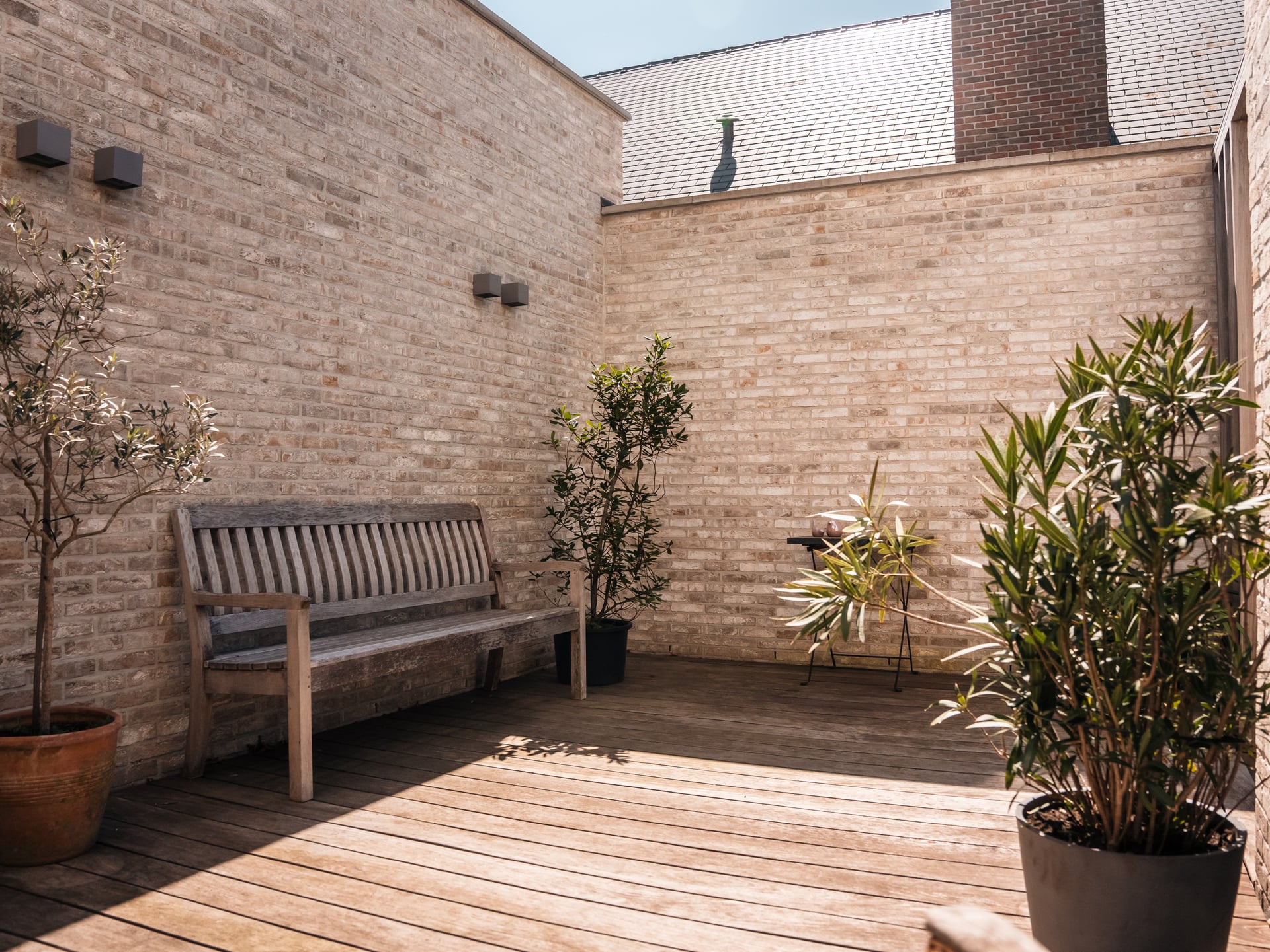 Zonnig houten terras met houten tuinbank en potplanten tegen een lichte bakstenen muur