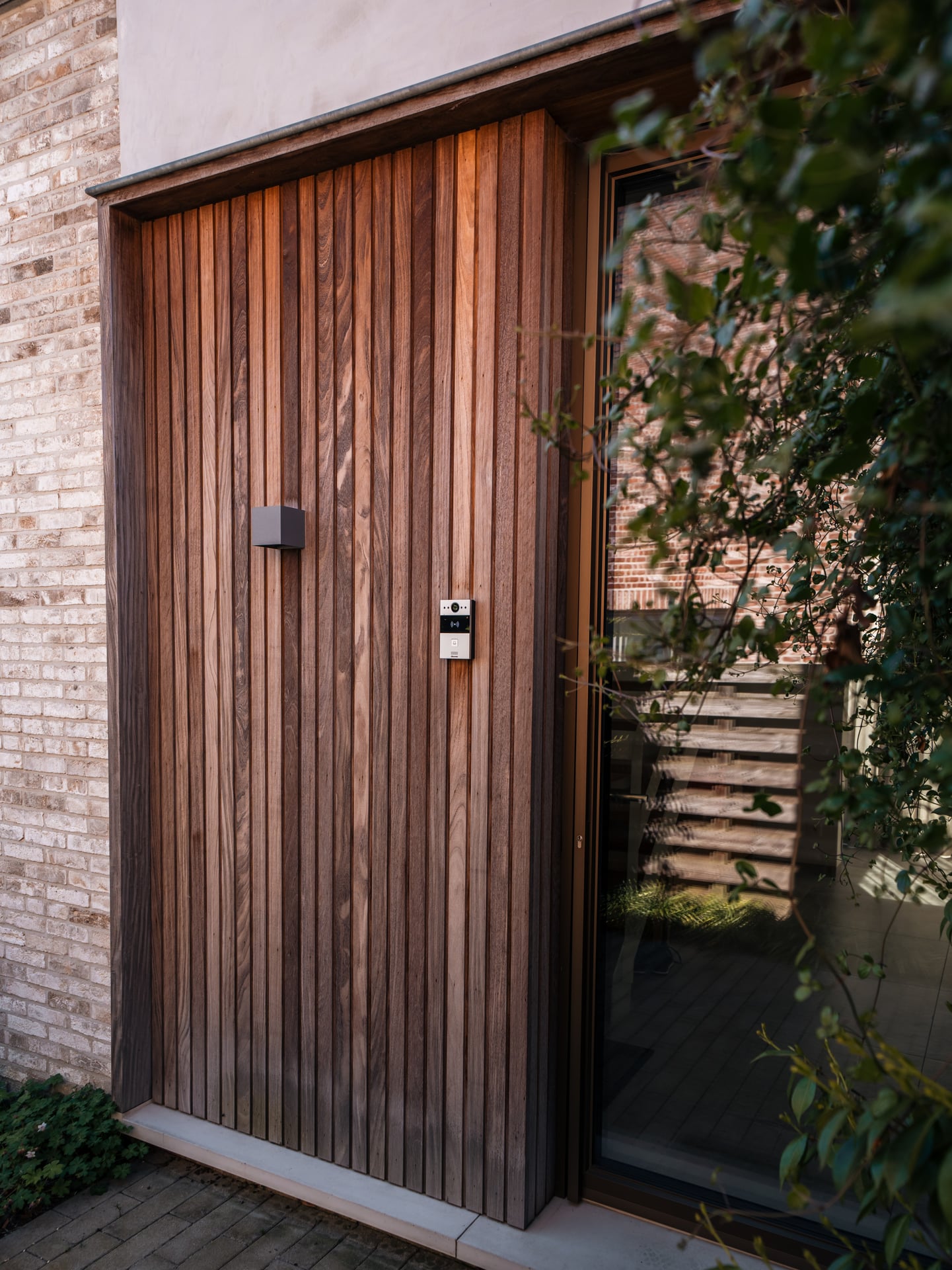 Voordeur met verticale houten latten in thermowood naast een glazen zijpaneel en klimop