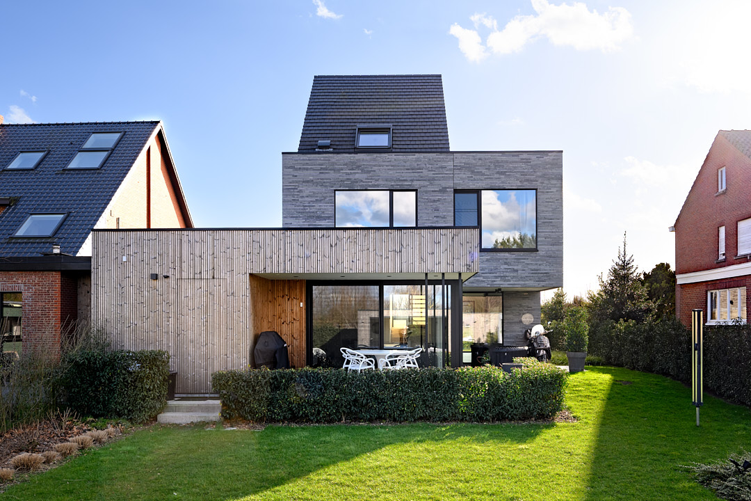 Achtergevel in Vremde met grijze gevelsteen, thermowood-aanbouw en overdekt terras met zwarte aluminium ramen