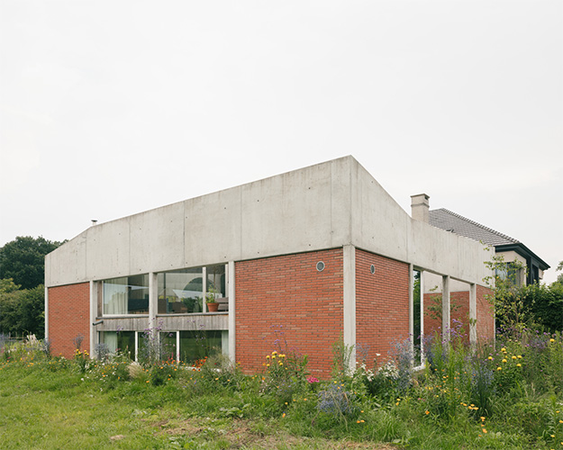 Hoekaanzicht van de betonnen woning met bakstenen invulling en omringende weide