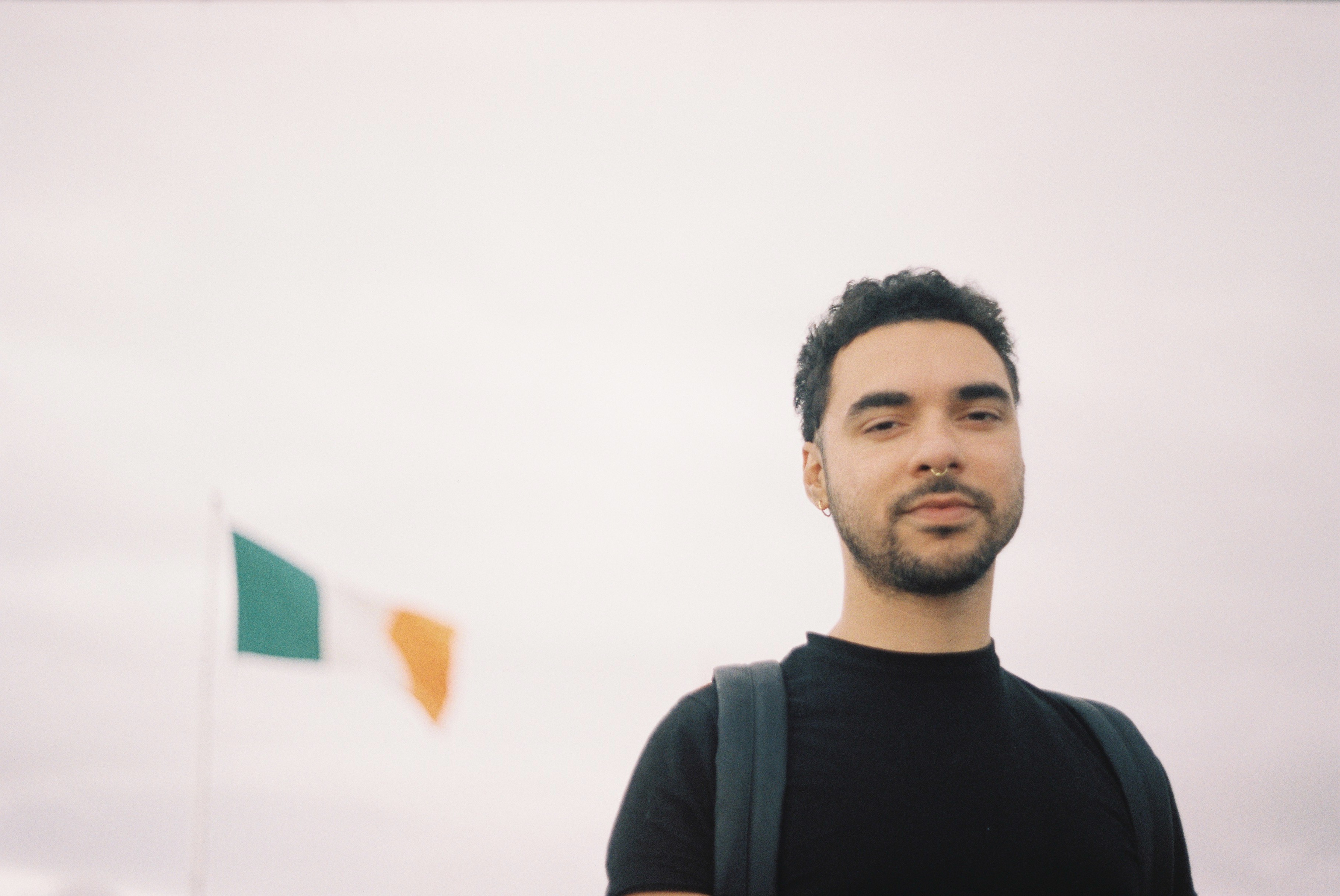 Young man with short curly hair and beard wearing a black shirt with an Irish flag in the background.