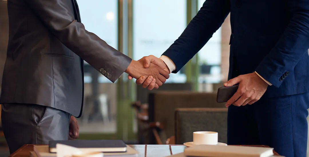 Two men in suits shaking hands over a table with notebooks and a cup in an office setting.