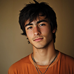 Young man with tousled dark hair, light facial hair, wearing an orange shirt and layered necklaces, looking at the camera with a slight smile.