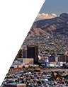 Cityscape with buildings and densely packed houses in front of mountainous terrain under a blue sky with a few clouds.