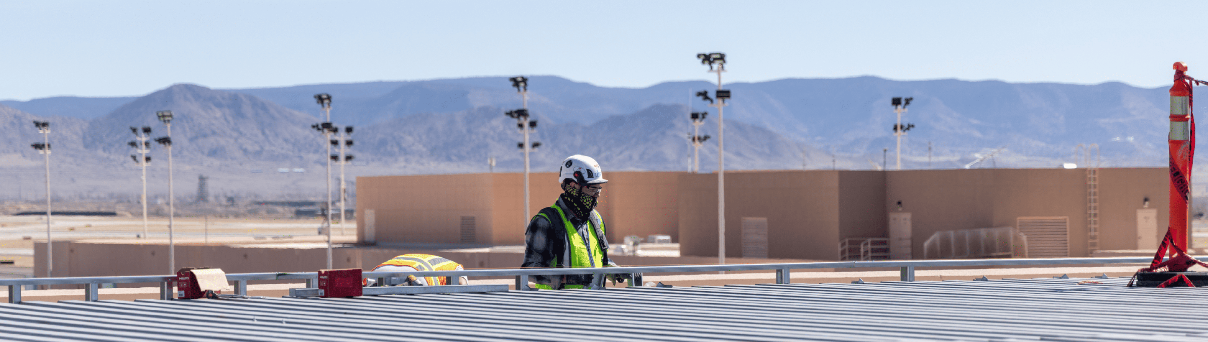 Construction worker wearing a helmet and safety vest working on a rooftop with mountains and industrial buildings in the background.