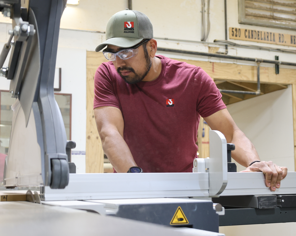 Man wearing safety glasses, a gray and green cap, and a maroon shirt with a logo, operating a woodworking table saw in a workshop.