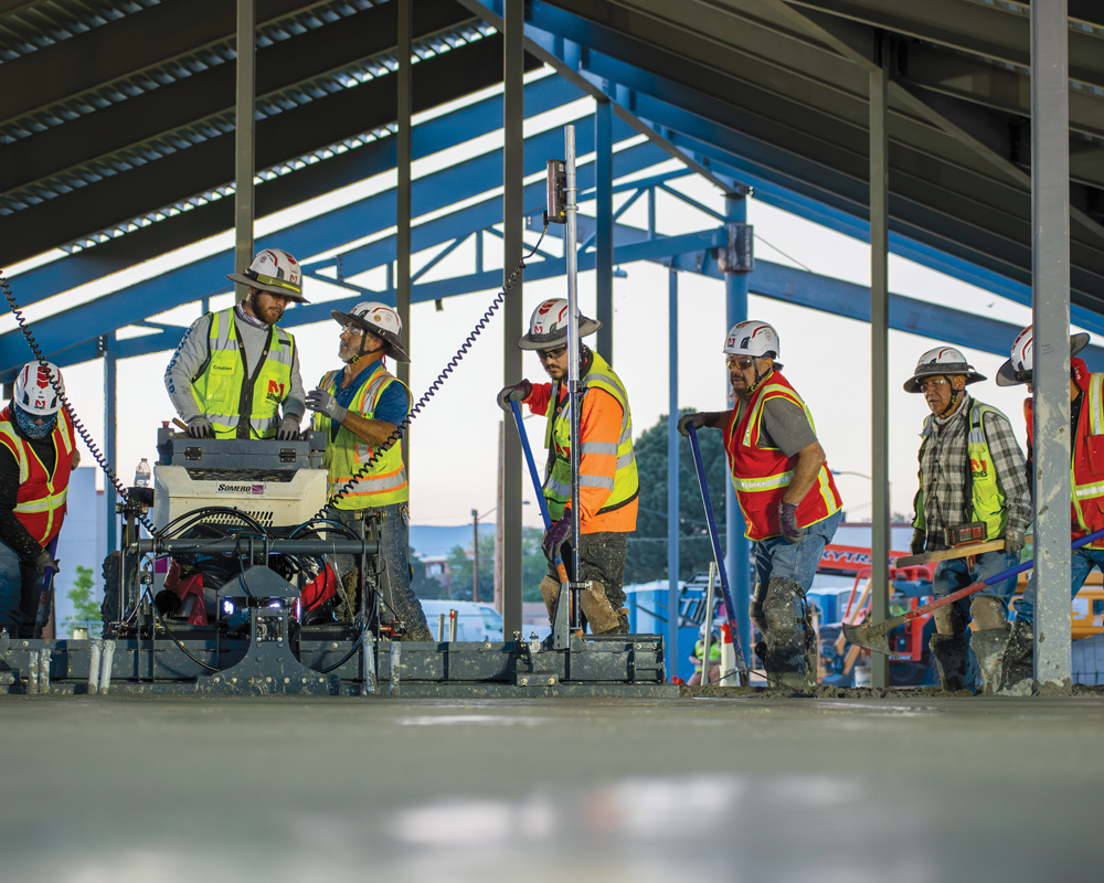 Construction workers wearing safety helmets and neon vests use tools and machinery to smooth and level freshly poured concrete inside a partially covered building frame.