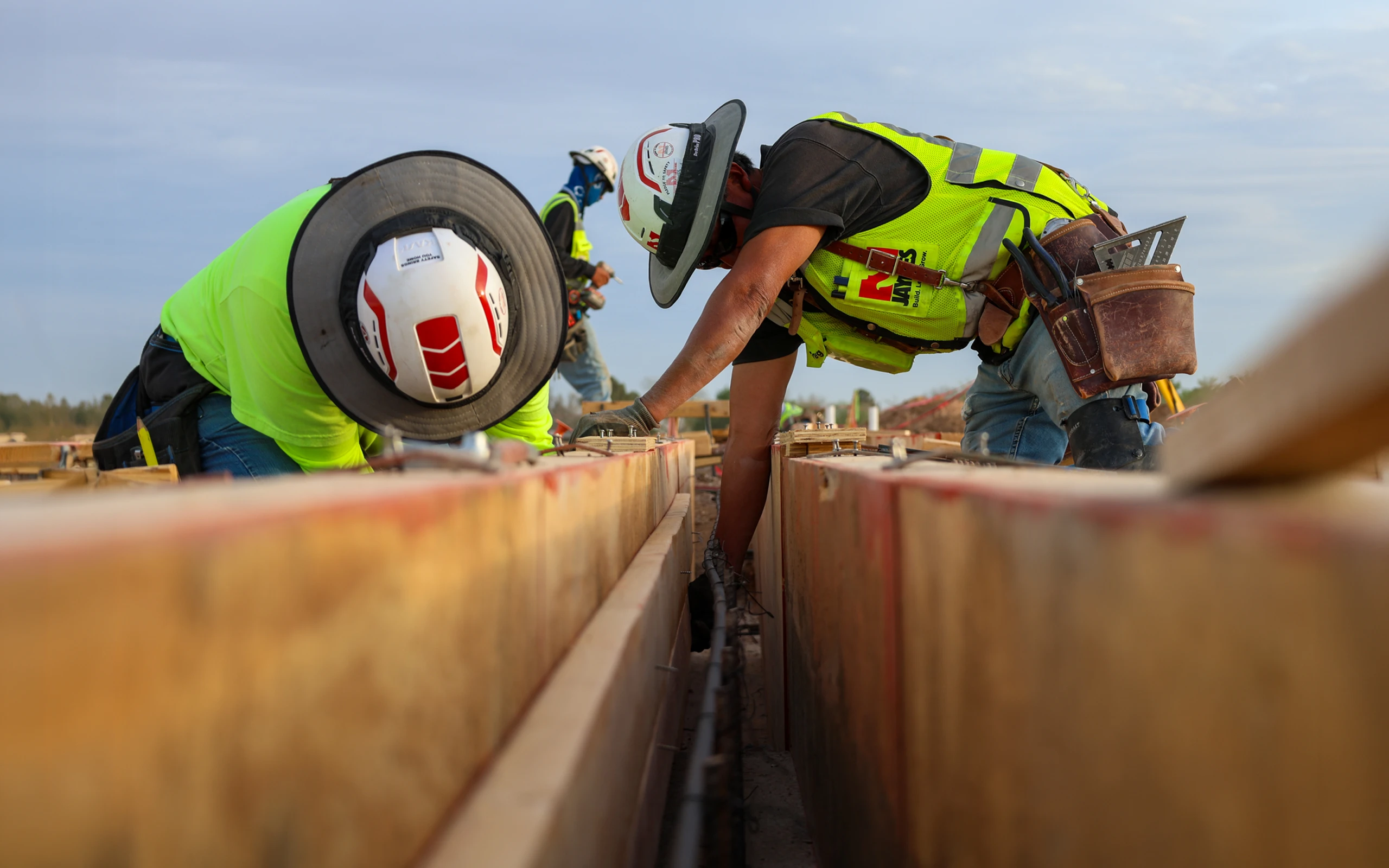 Three construction workers in safety vests and white helmets working on a wooden structure, fixing wiring or piping inside a narrow trench between wooden boards at an outdoor site under a clear sky.