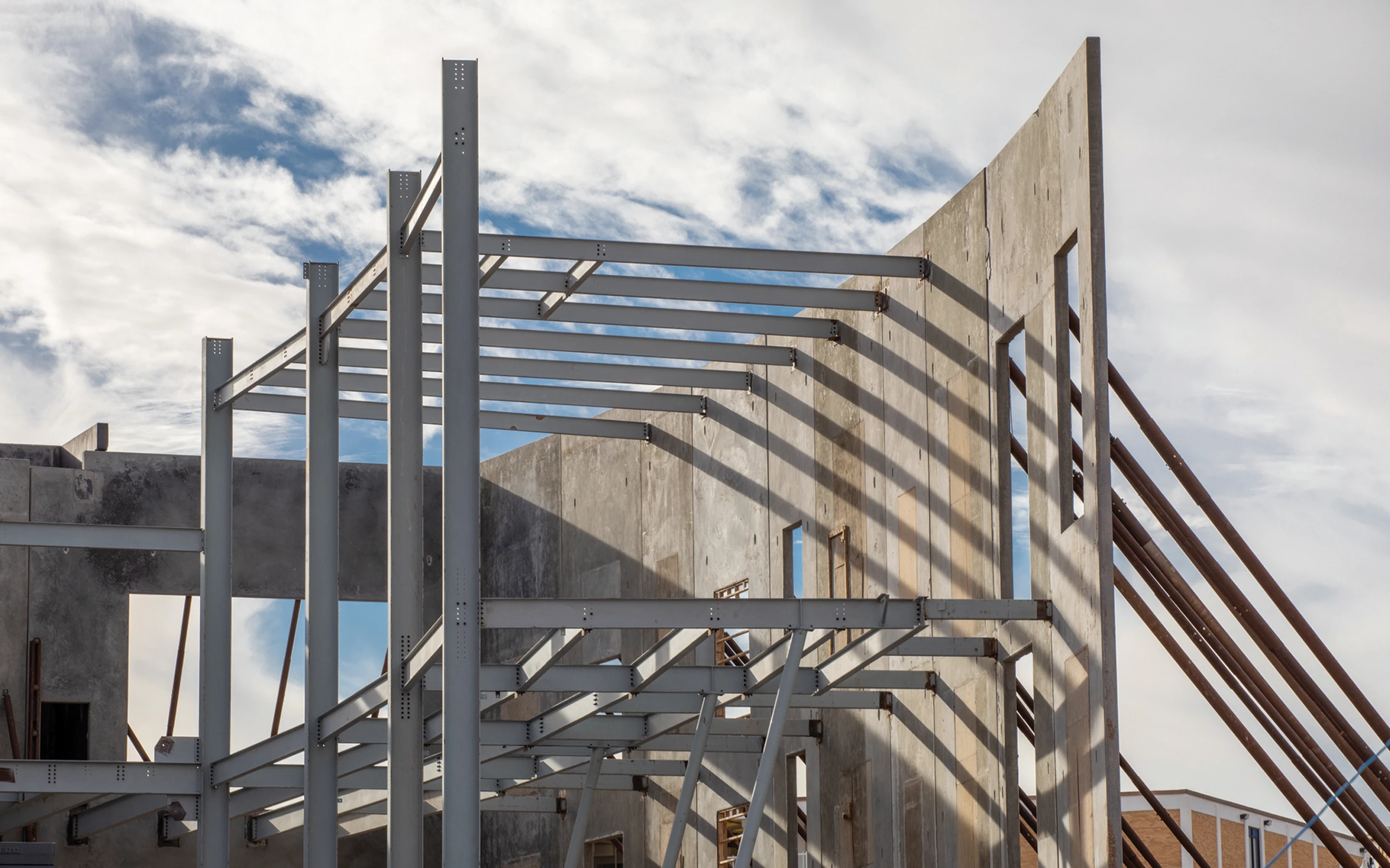 Partially constructed building showing concrete walls supported by diagonal steel braces and a steel framework extending above, with blue sky and clouds in the background.