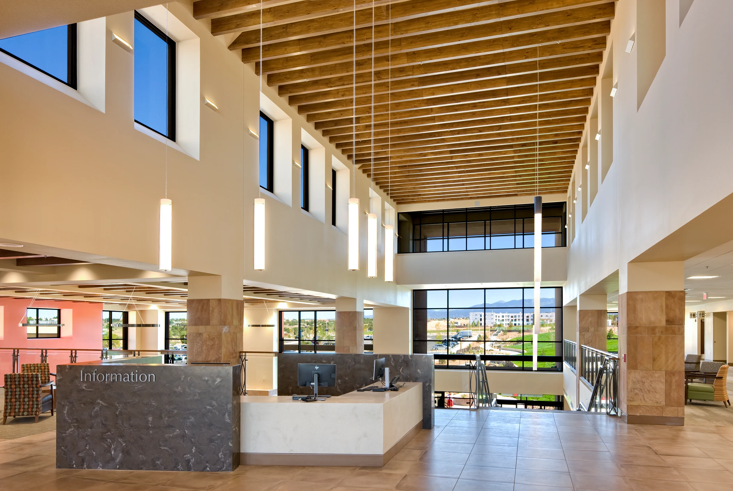 Spacious, modern lobby area with high wood-beamed ceiling, vertical hanging lights, large windows with mountain view, and an information desk with two computer monitors.
