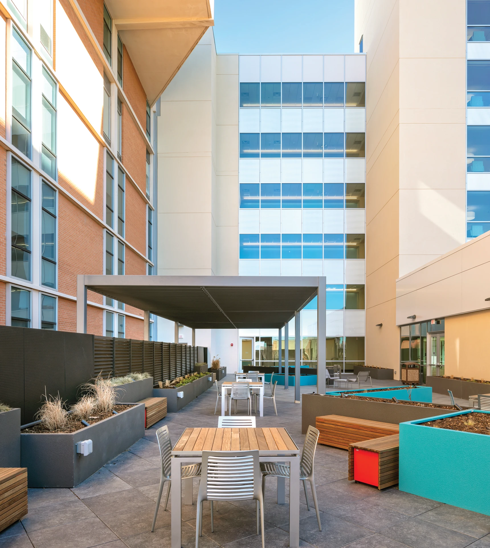 Modern outdoor seating area between buildings with tables, chairs, planter boxes with dry plants, and a shaded pergola under a clear sky.