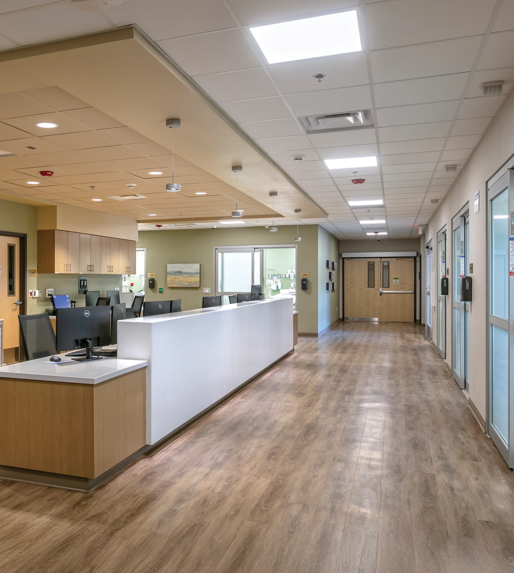 Clean, modern hospital nursing station with multiple computer monitors on the left and a long corridor with patient rooms and closed wooden double doors at the end.