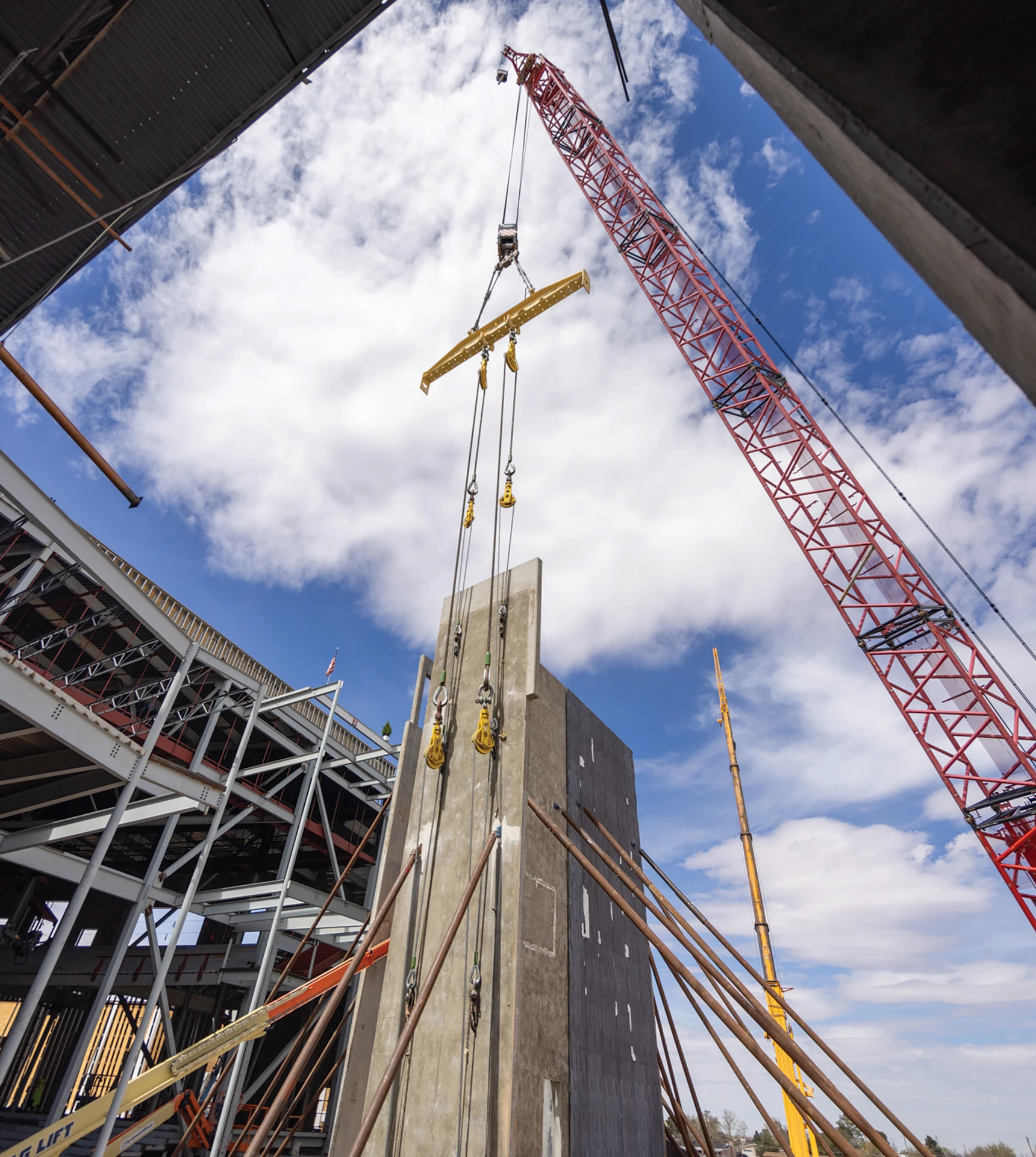 Red crane lifting a large concrete wall panel at a construction site with steel framework and support beams under a partly cloudy sky.