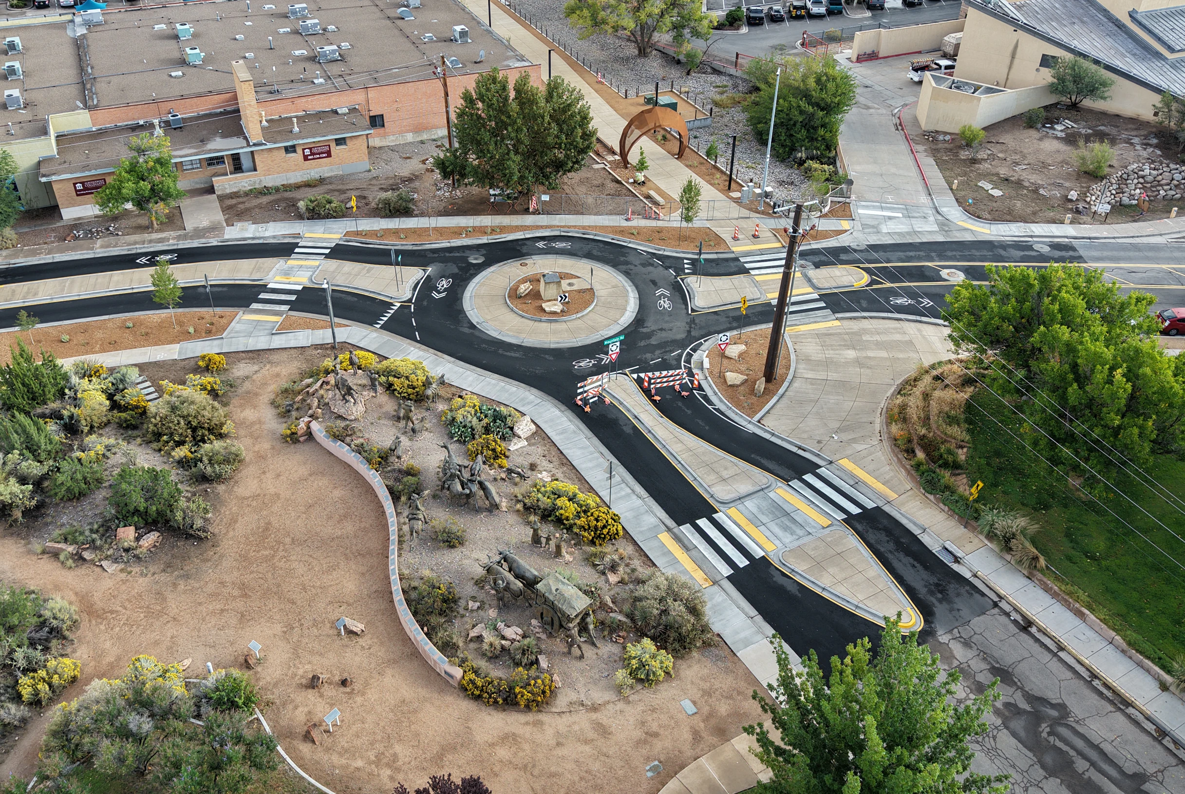 Aerial view of a small roundabout with bike lanes and pedestrian crosswalks near a road and a building area, including landscaped patches with shrubs and a bronze sculpture exhibit featuring oxen and people.