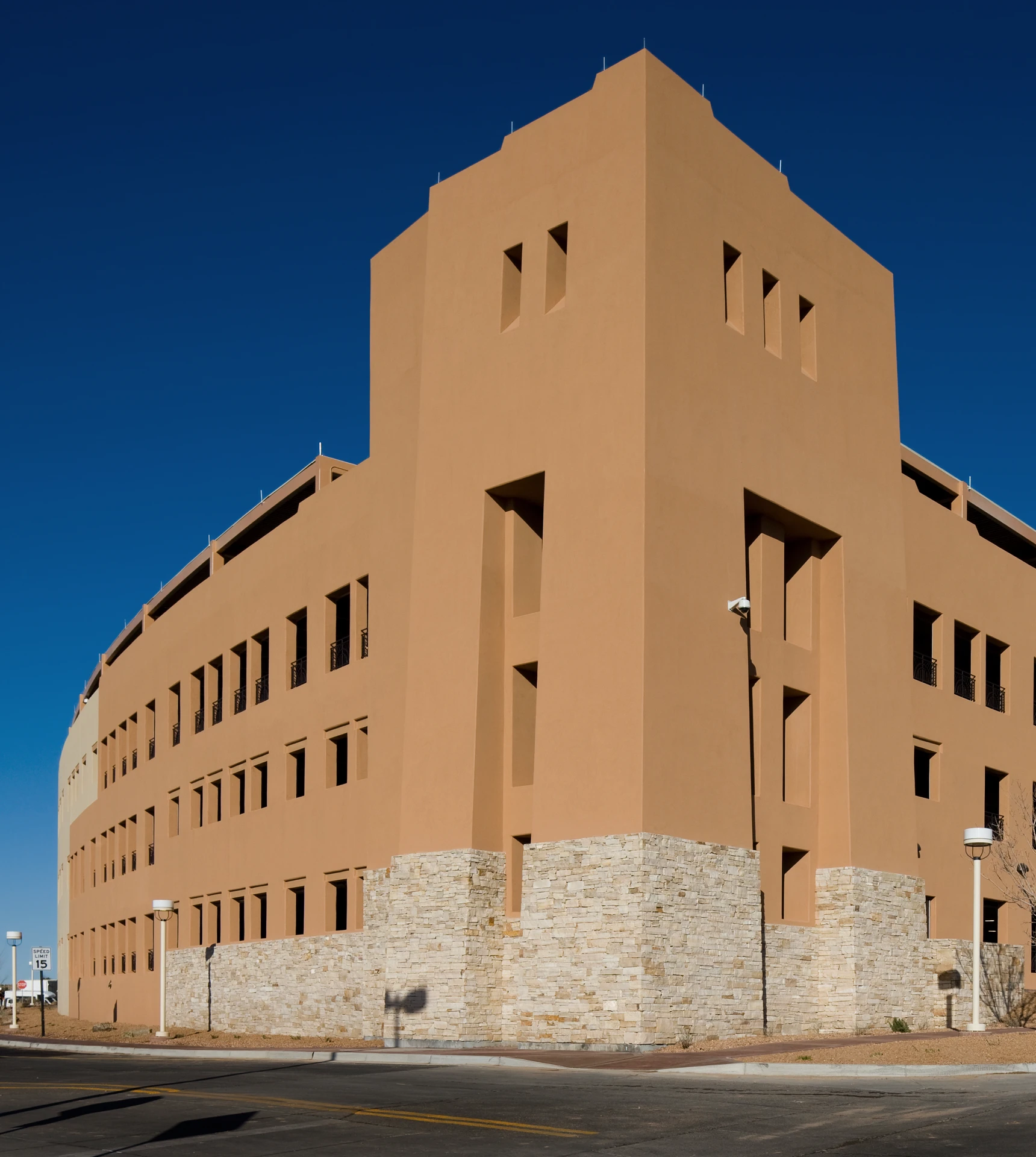 Large beige stucco building with multiple rectangular recessed windows and a lower stone wall base, seen against a clear deep blue sky on a sunny day, located at a street corner with street lamps and a speed limit sign.