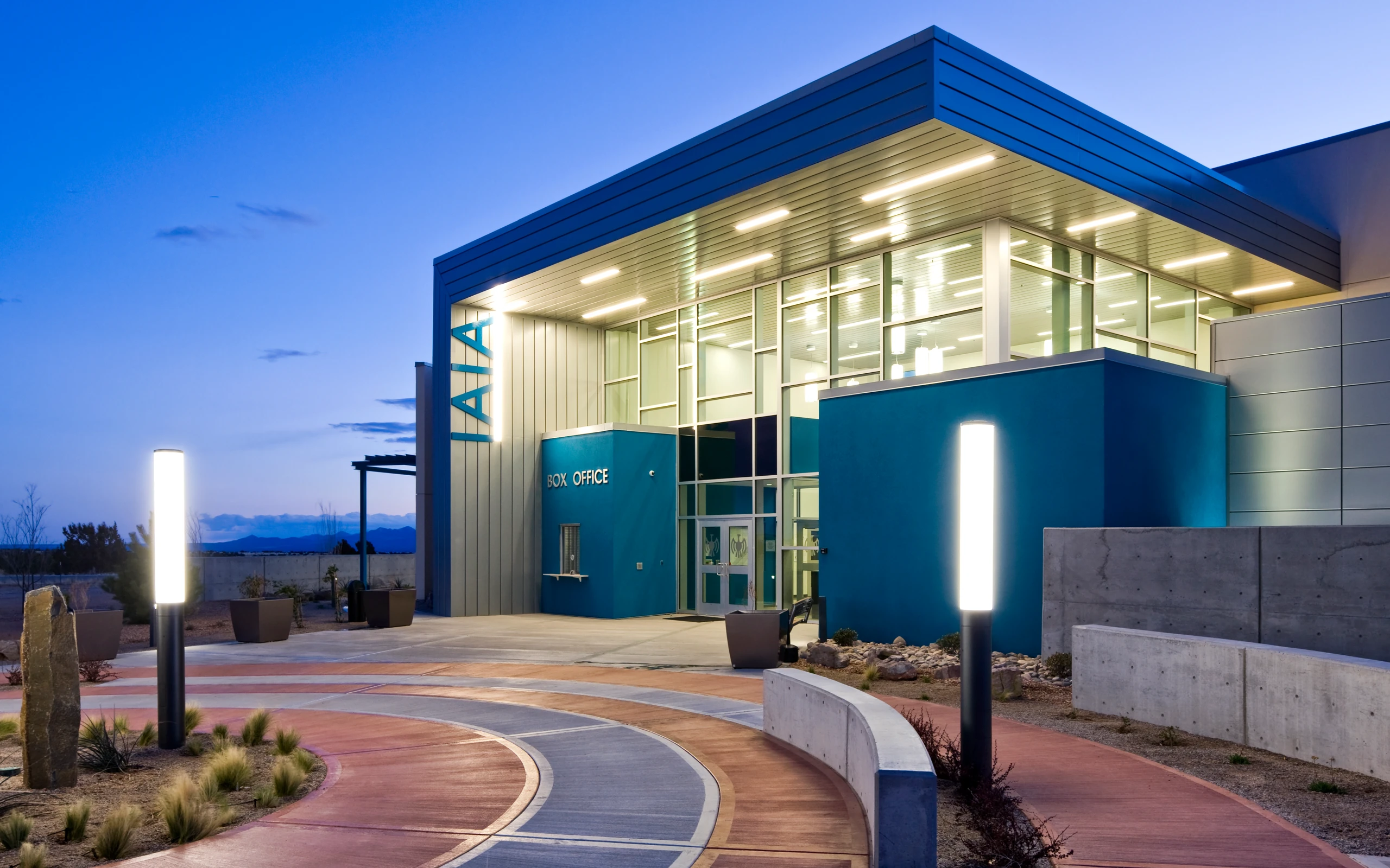 Modern performing arts center building at dusk with large glass windows, blue and gray exterior walls, illuminated overhead lights, a box office window, curved concrete pathways, and tall glowing light posts.