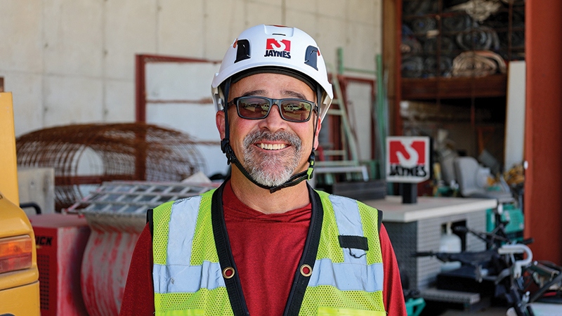 Smiling construction worker wearing a white Jaynes hard hat, reflective yellow safety vest, red shirt, and glasses, standing in front of construction materials and equipment.