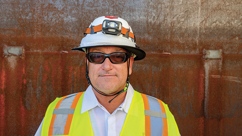 Man wearing safety gear including a white hard hat with a mounted headlamp, dark safety glasses, and a high-visibility yellow and orange reflective vest, standing against a rusted metal background.